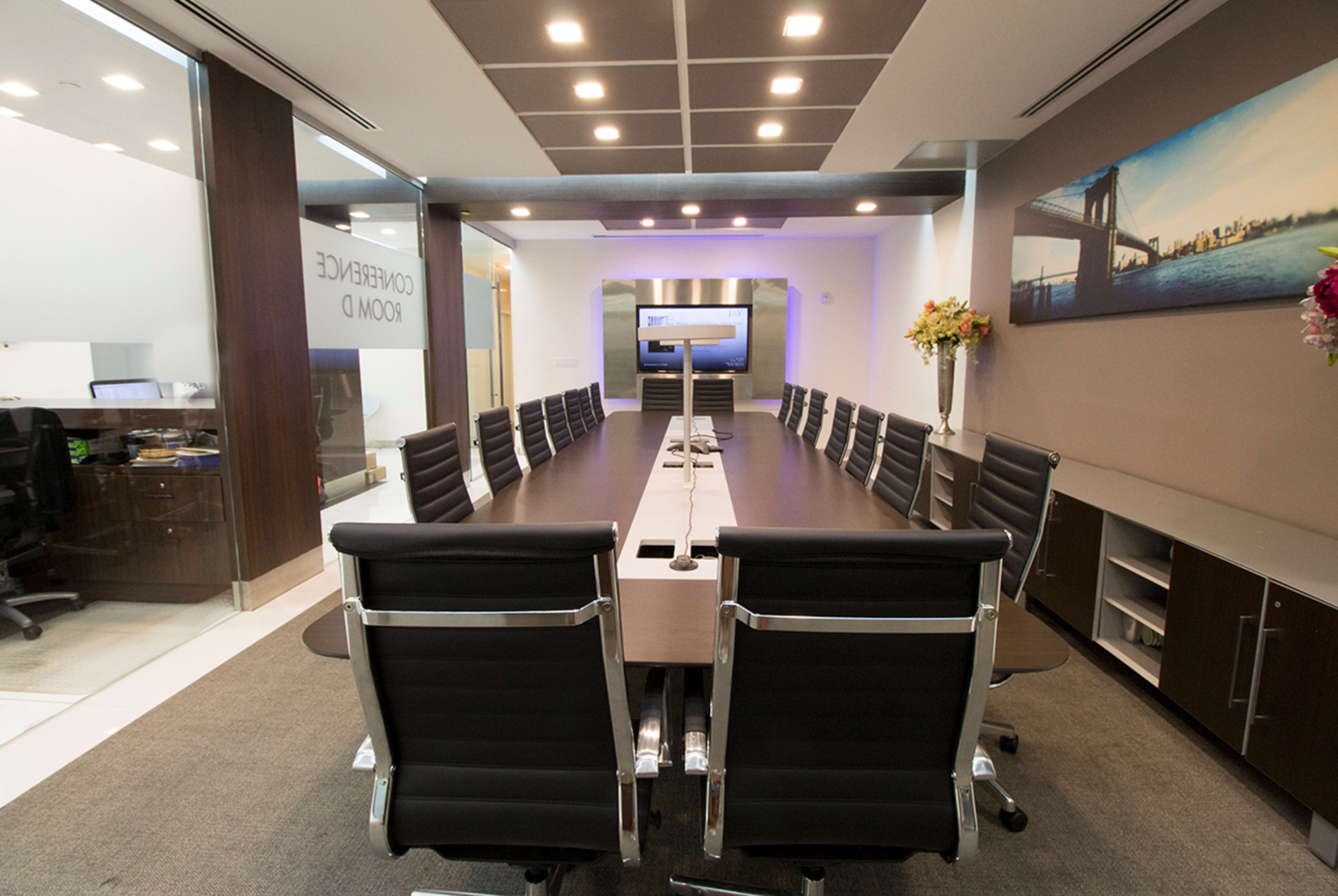 A long conference table with black leather chairs is set in a modern meeting room featuring a large screen and a photo of the Brooklyn Bridge on the wall.