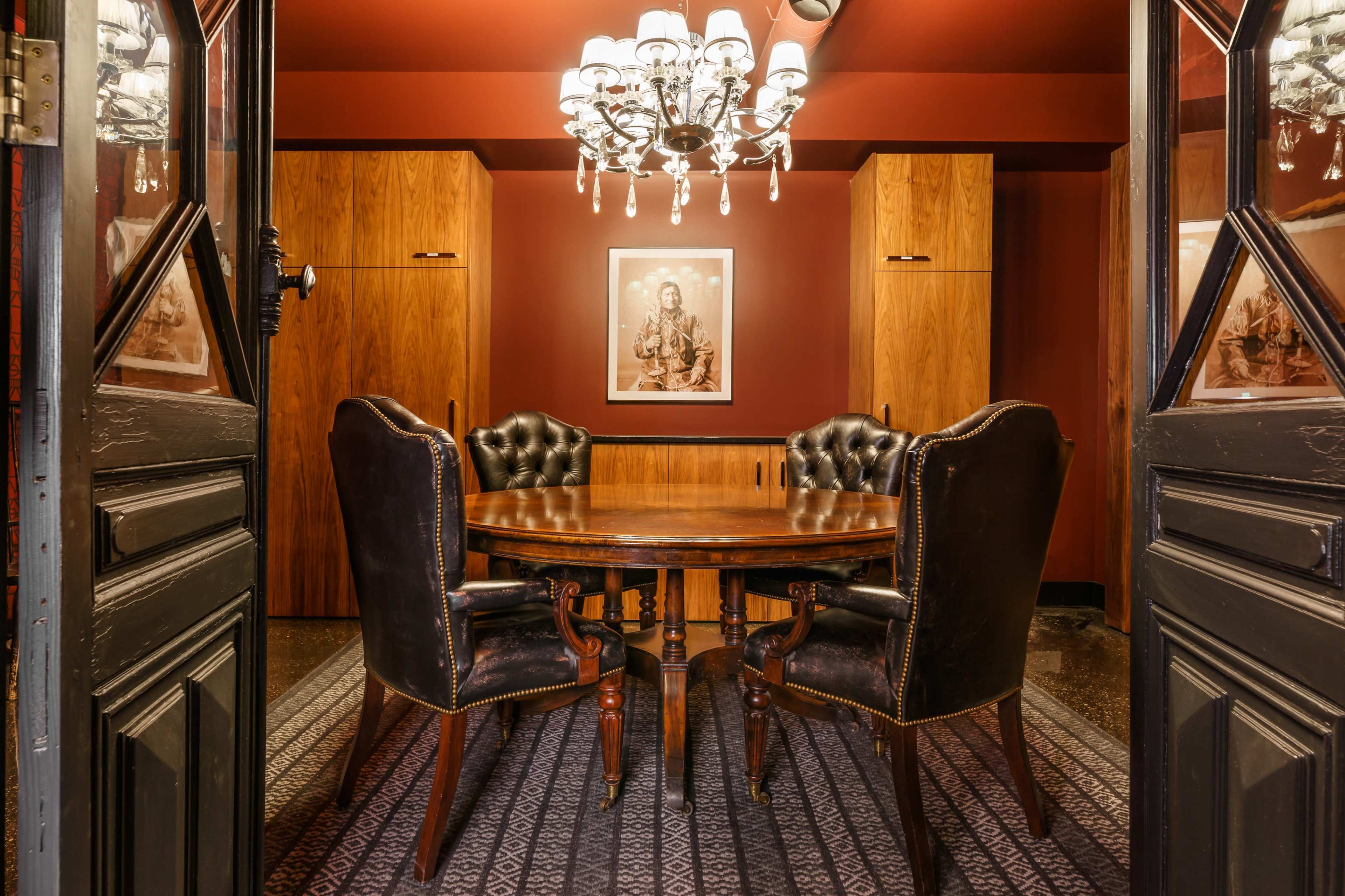A wooden dining table with four black upholstered chairs is set in a dimly lit room adorned with dark red walls and a chandelier.