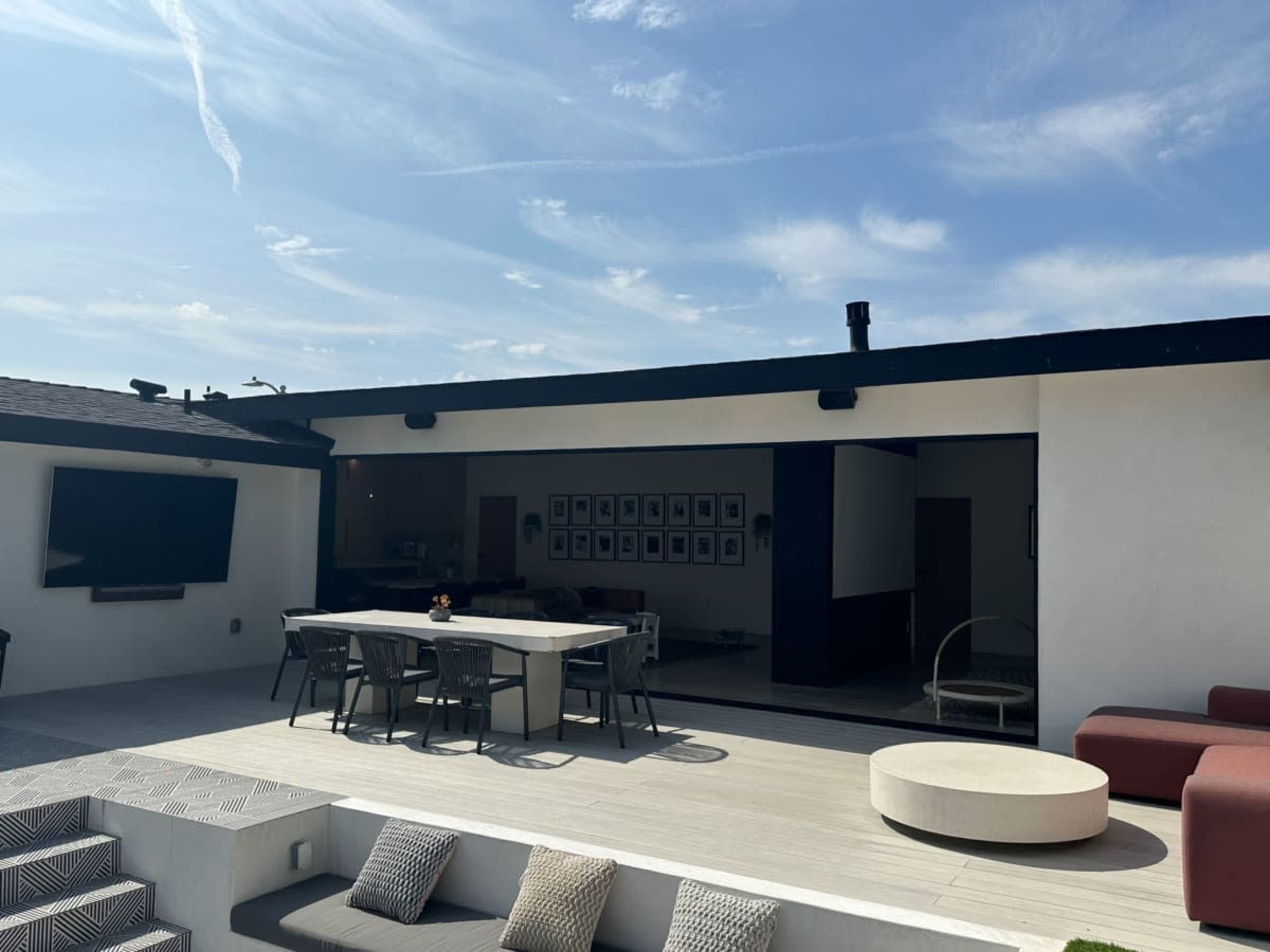 The image shows a modern outdoor patio area featuring a dining table, lounge seating, and an adjacent indoor space with a large opening.