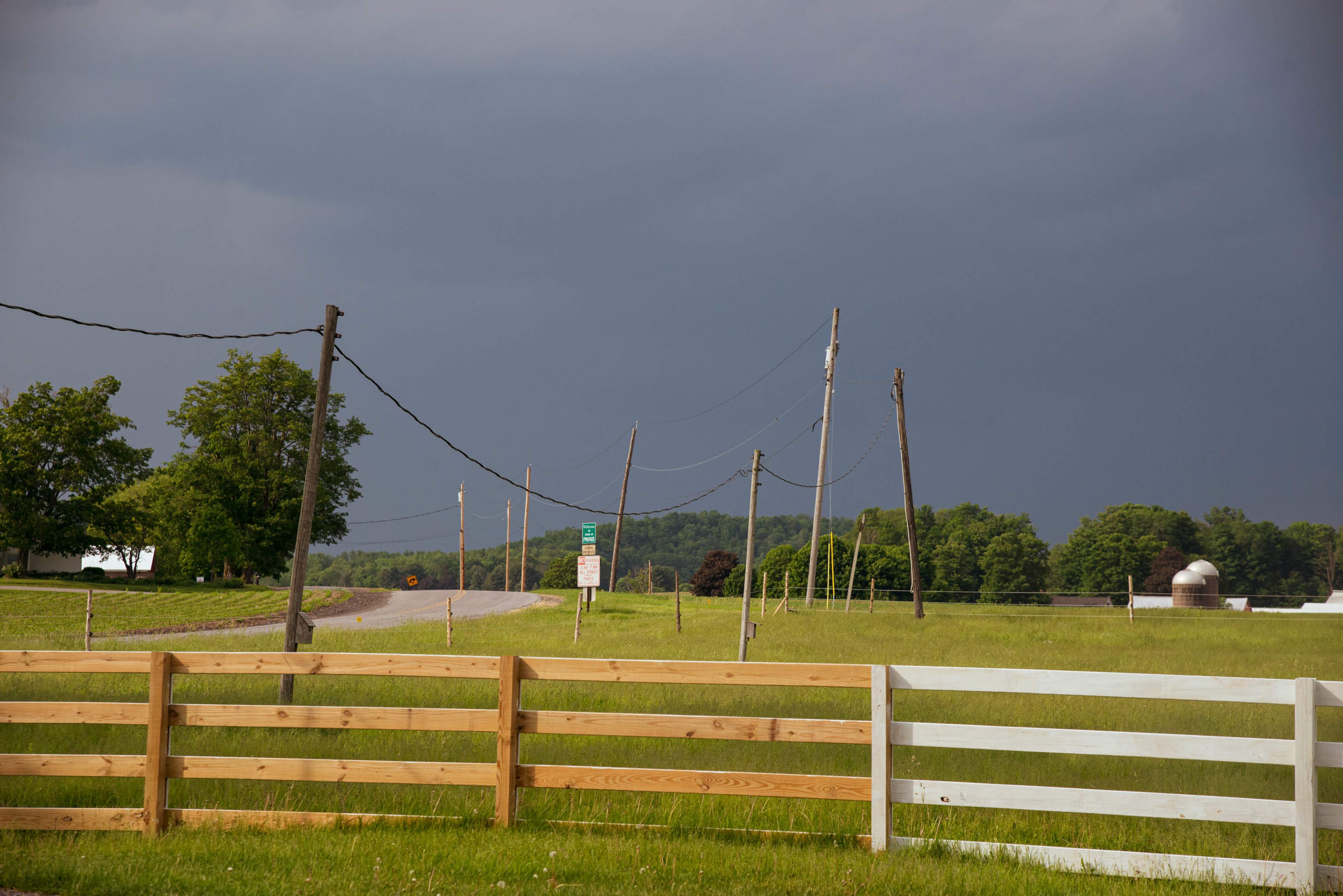 A dirt road runs through an open field with a wooden fence in the foreground and dark storm clouds looming in the background.