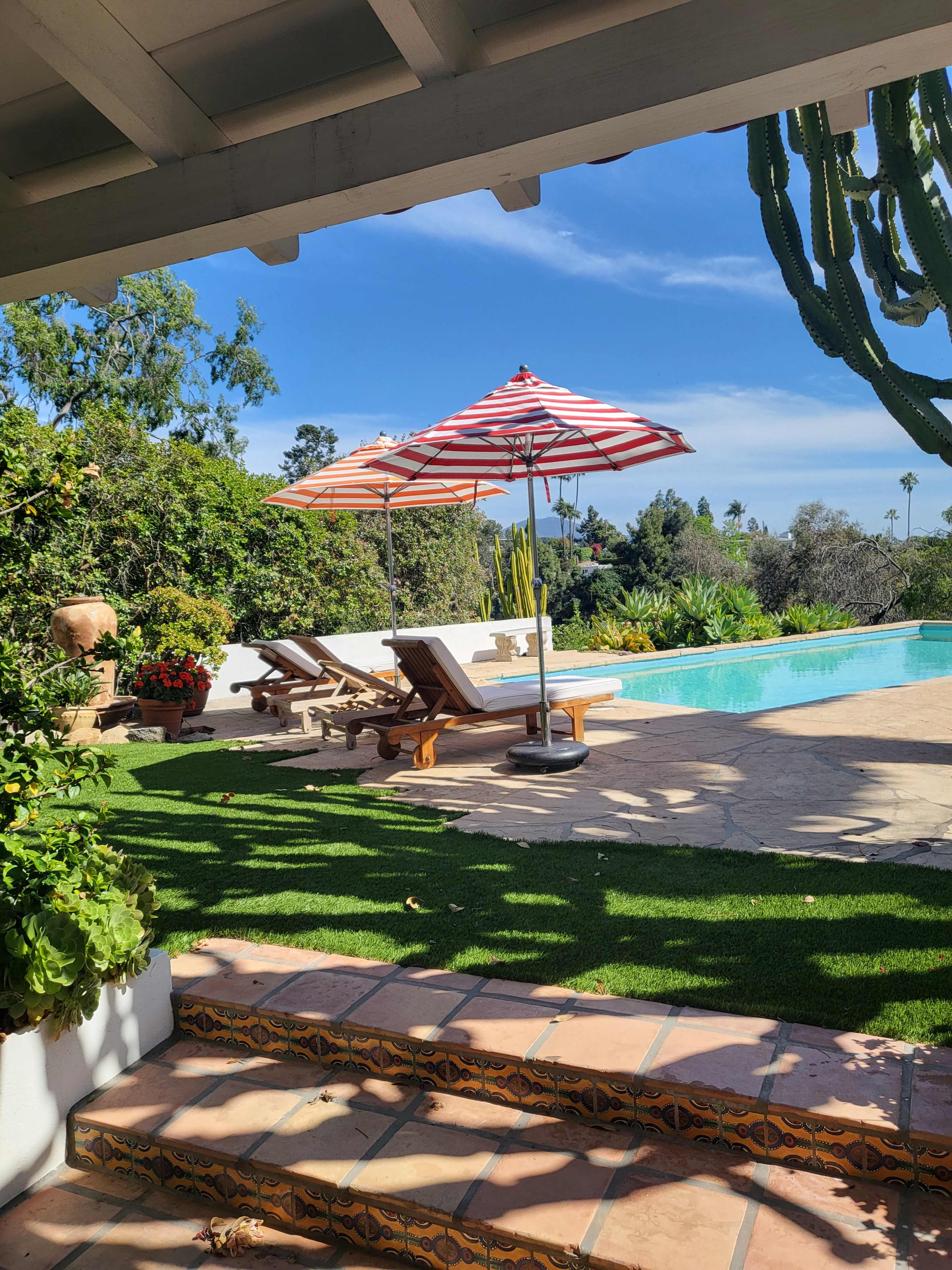 A landscaped backyard with a swimming pool, two lounge chairs under striped umbrellas, and steps leading up from a tiled patio.