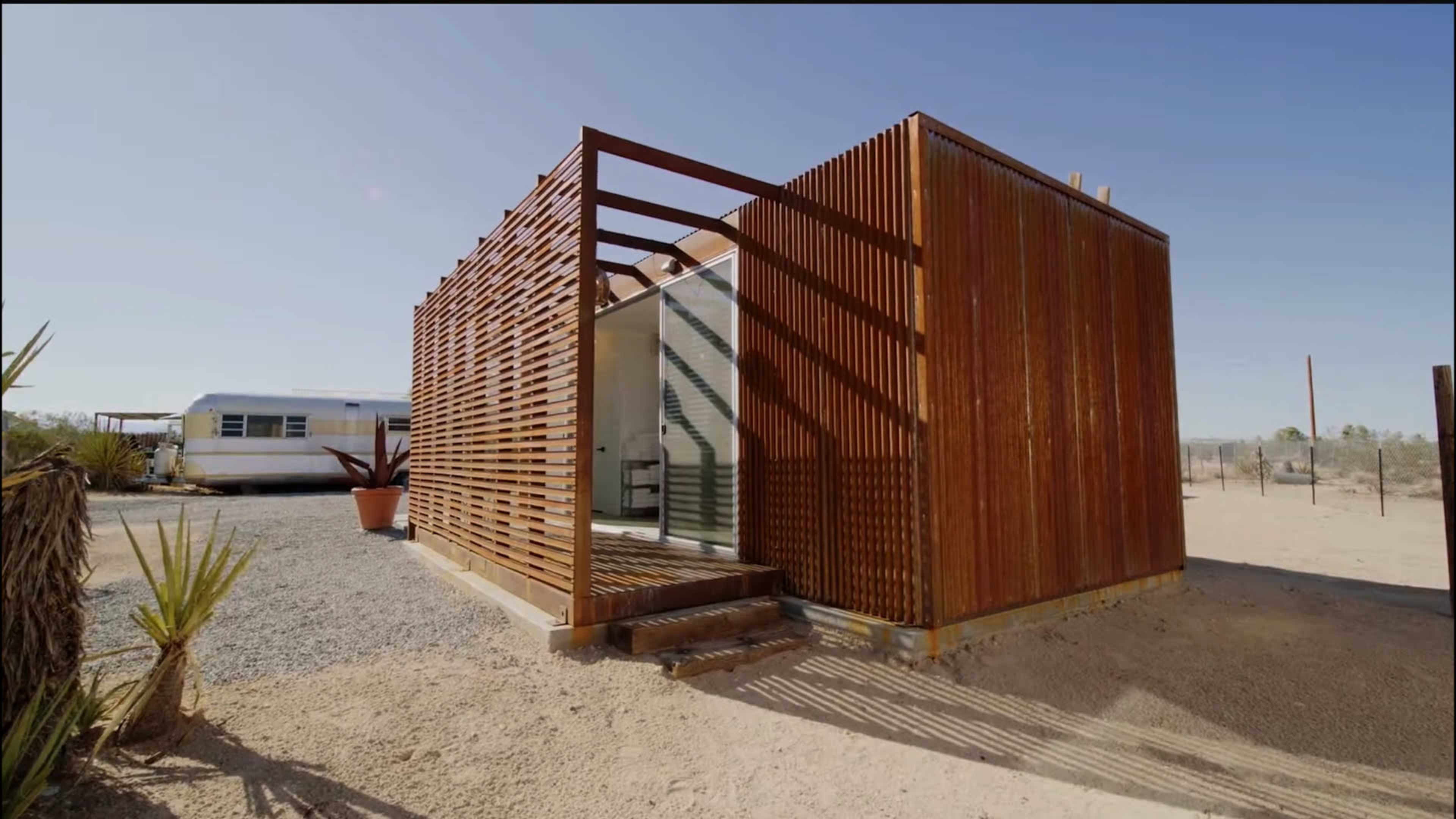 The image shows a modern, wooden slatted cabin set in a desert landscape, with a clear blue sky in the background.