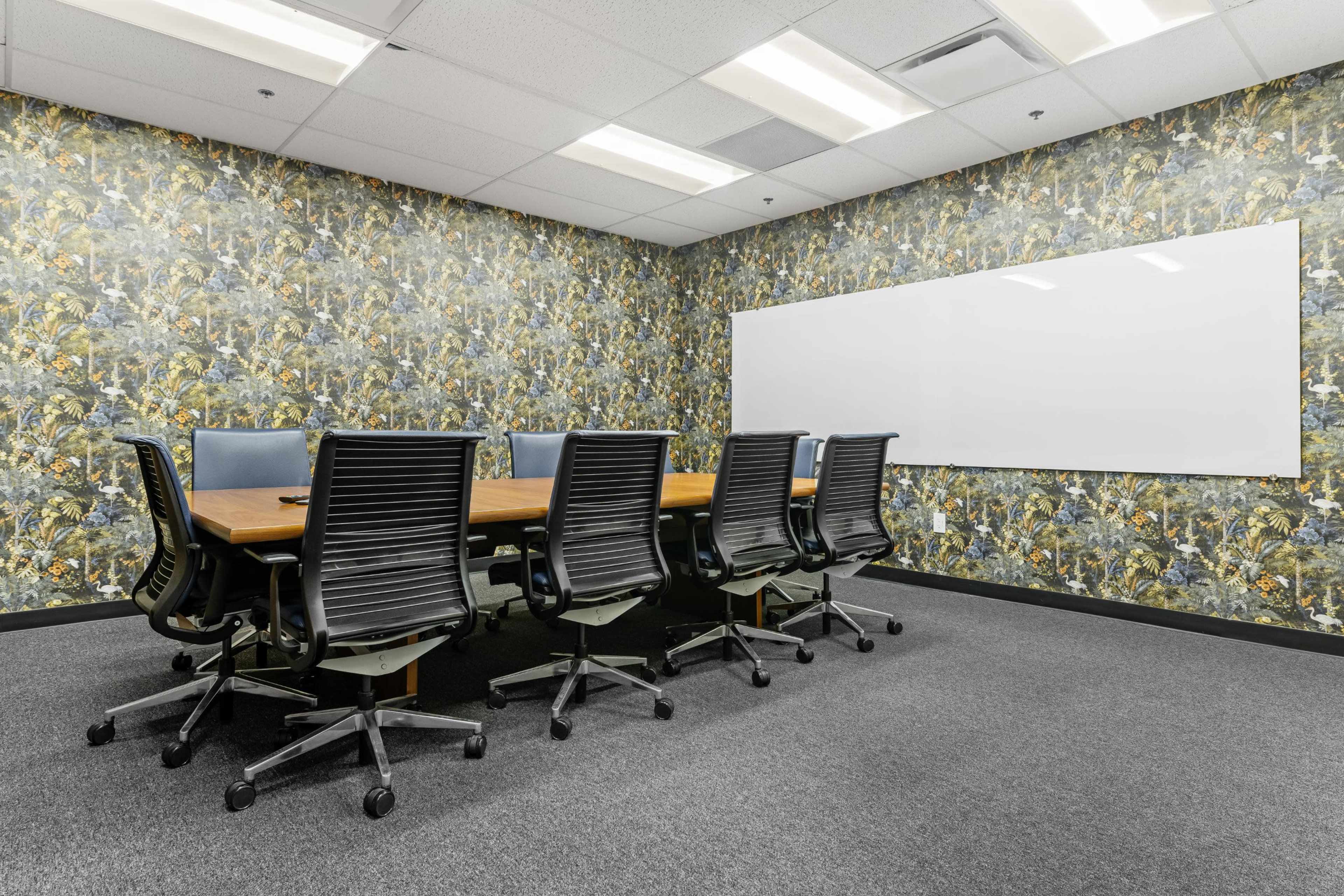 A conference room features a long wooden table surrounded by six black rolling chairs, with a floral-patterned wallpaper and a whiteboard on one wall.
