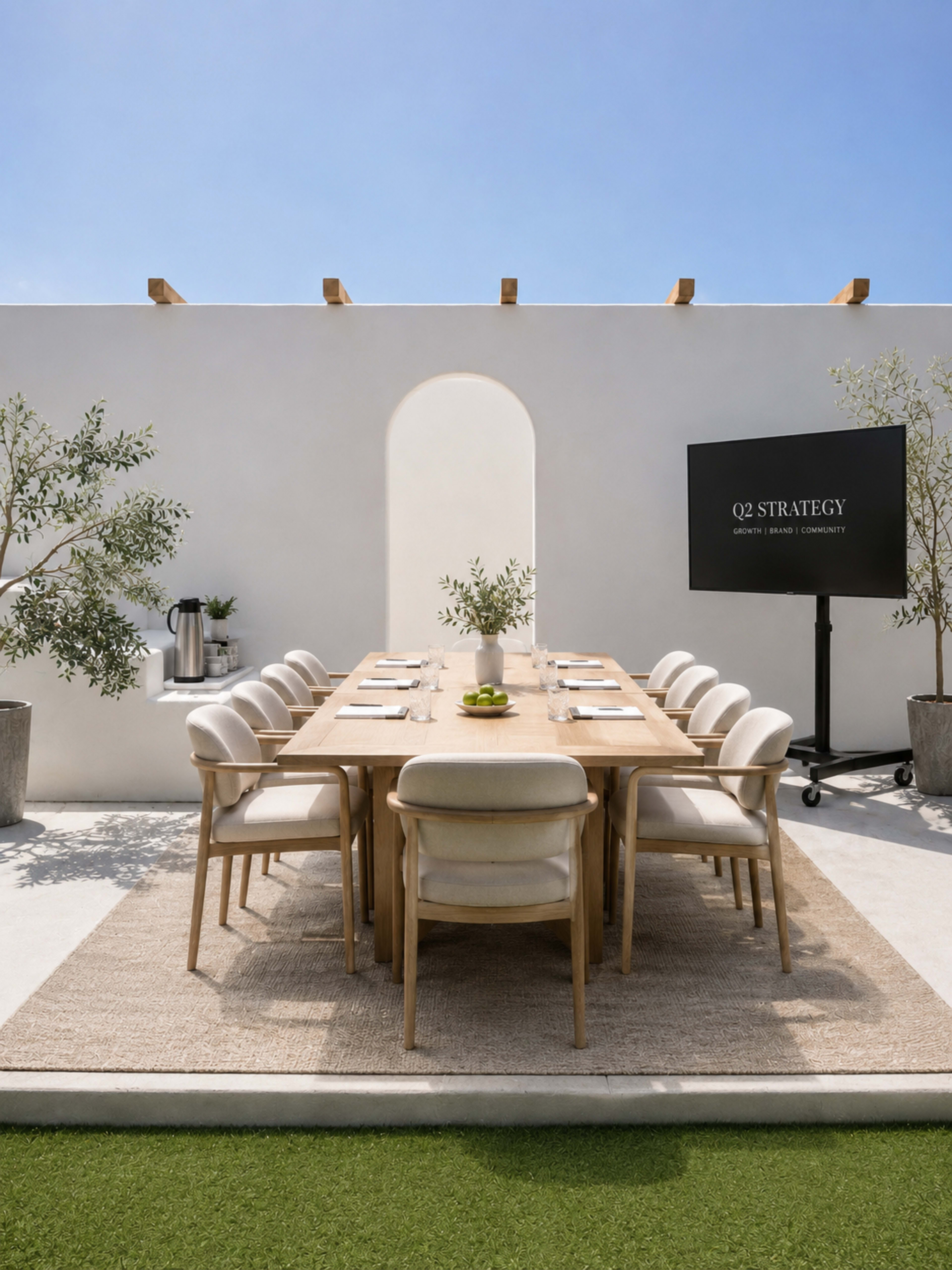 A long wooden dining table with white chairs is set up outdoors under a blue sky, accompanied by greenery and a presentation screen displaying "Q2 STRATEGY."