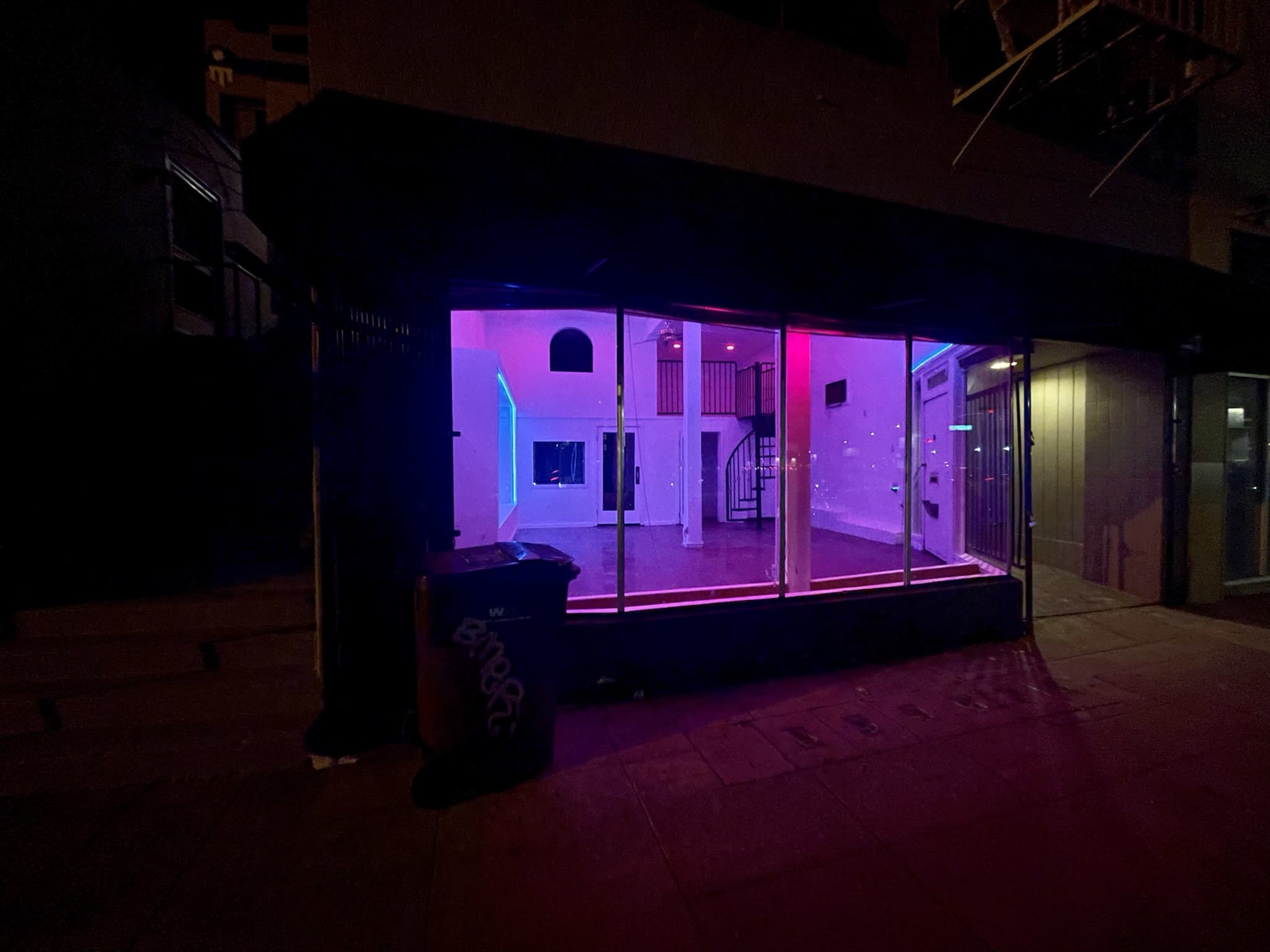 A dimly lit storefront features vibrant pink and blue lighting with an interior showcasing an open space and staircase.