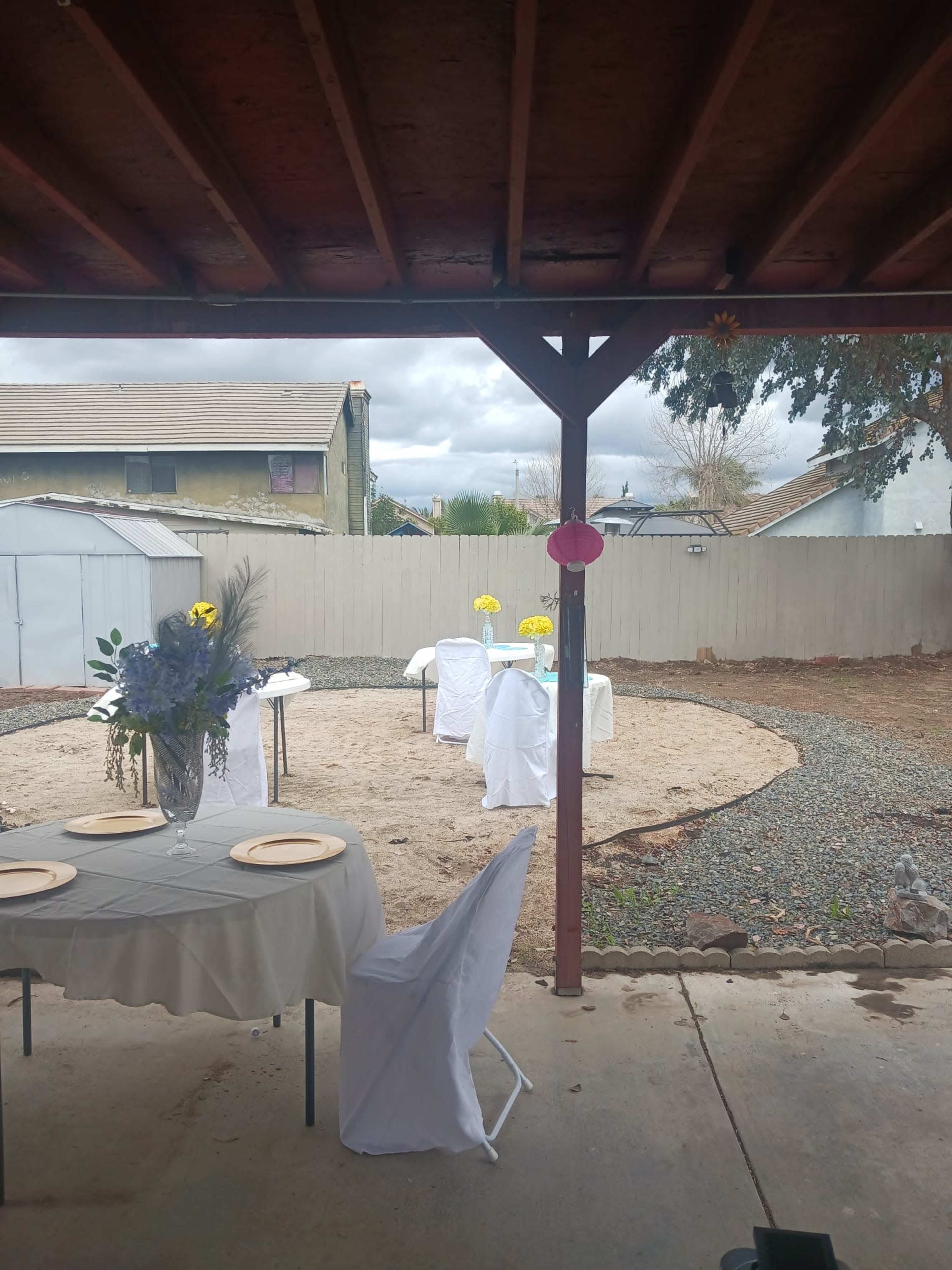 An outdoor patio area features a dining table set with plates and a vase of flowers, overlooking a gravel courtyard with white chairs and table arrangements.