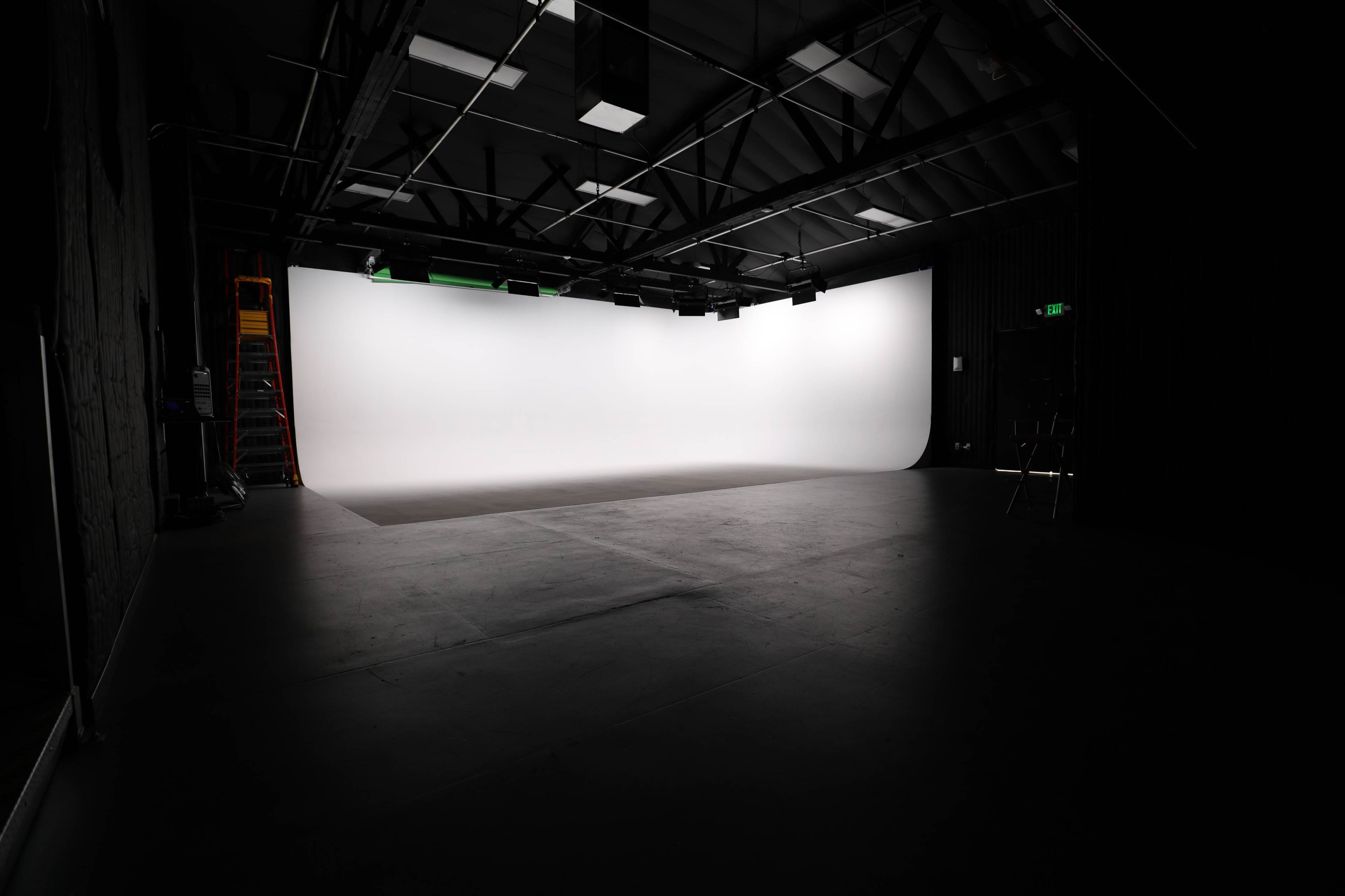 The image shows a dimly lit studio space featuring a large white backdrop, a few overhead lights, and a ladder against the wall.