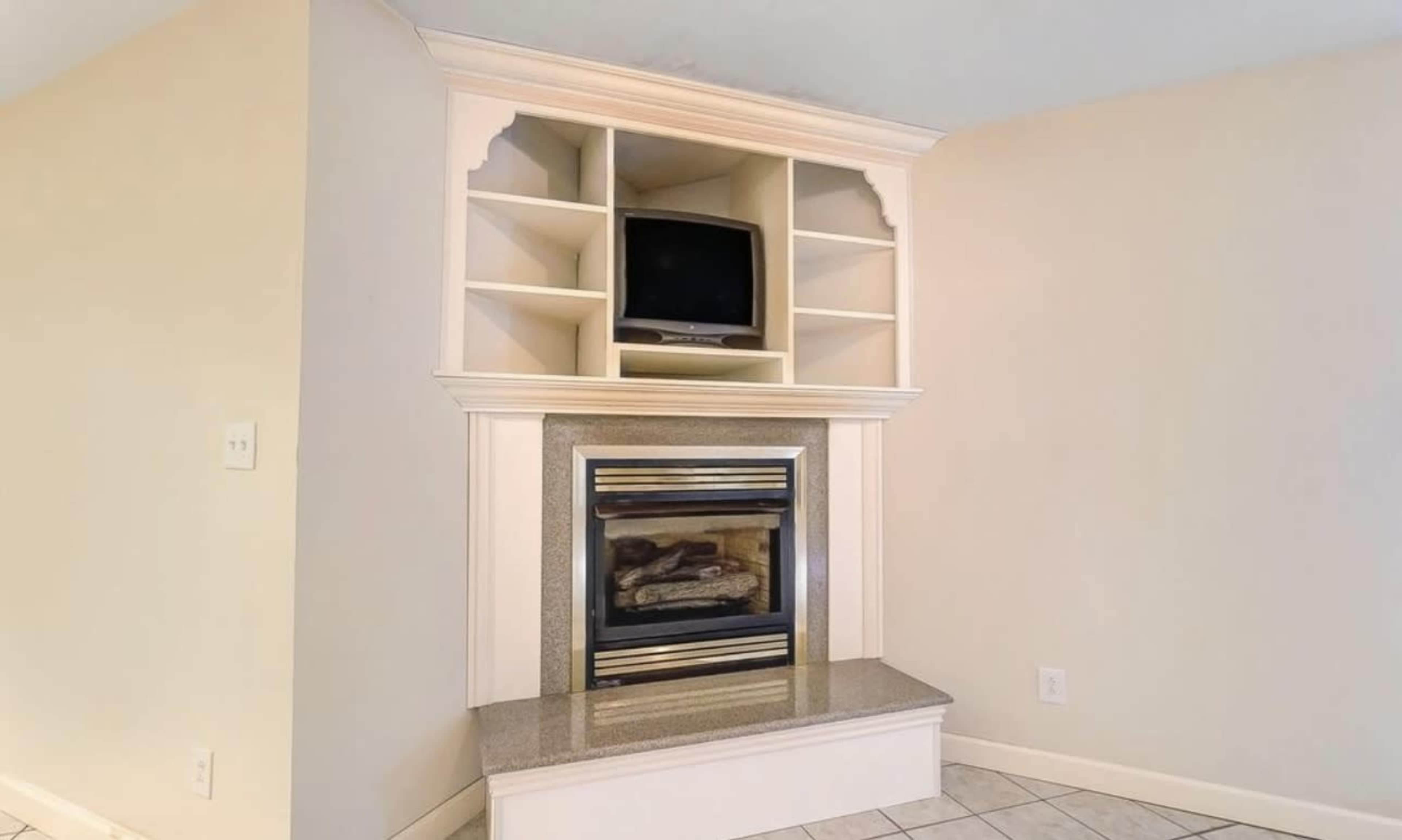 The image shows a corner fireplace with a built-in television shelf above it, surrounded by light-colored walls and tile flooring.