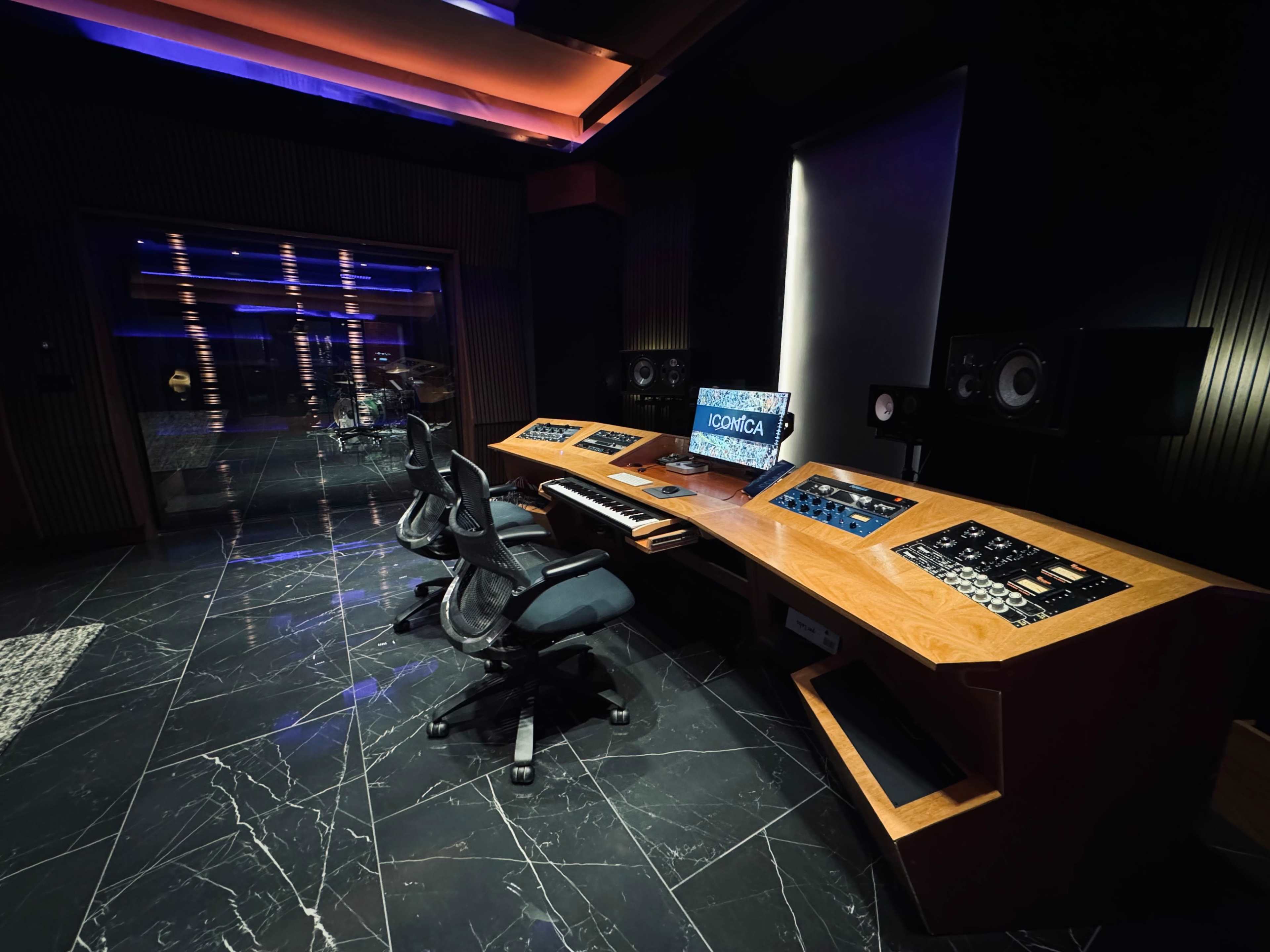 A modern music studio with a large wooden console, three black chairs, and a computer monitor displaying "ICMICA" against a backdrop of dark marble flooring and ambient lighting.