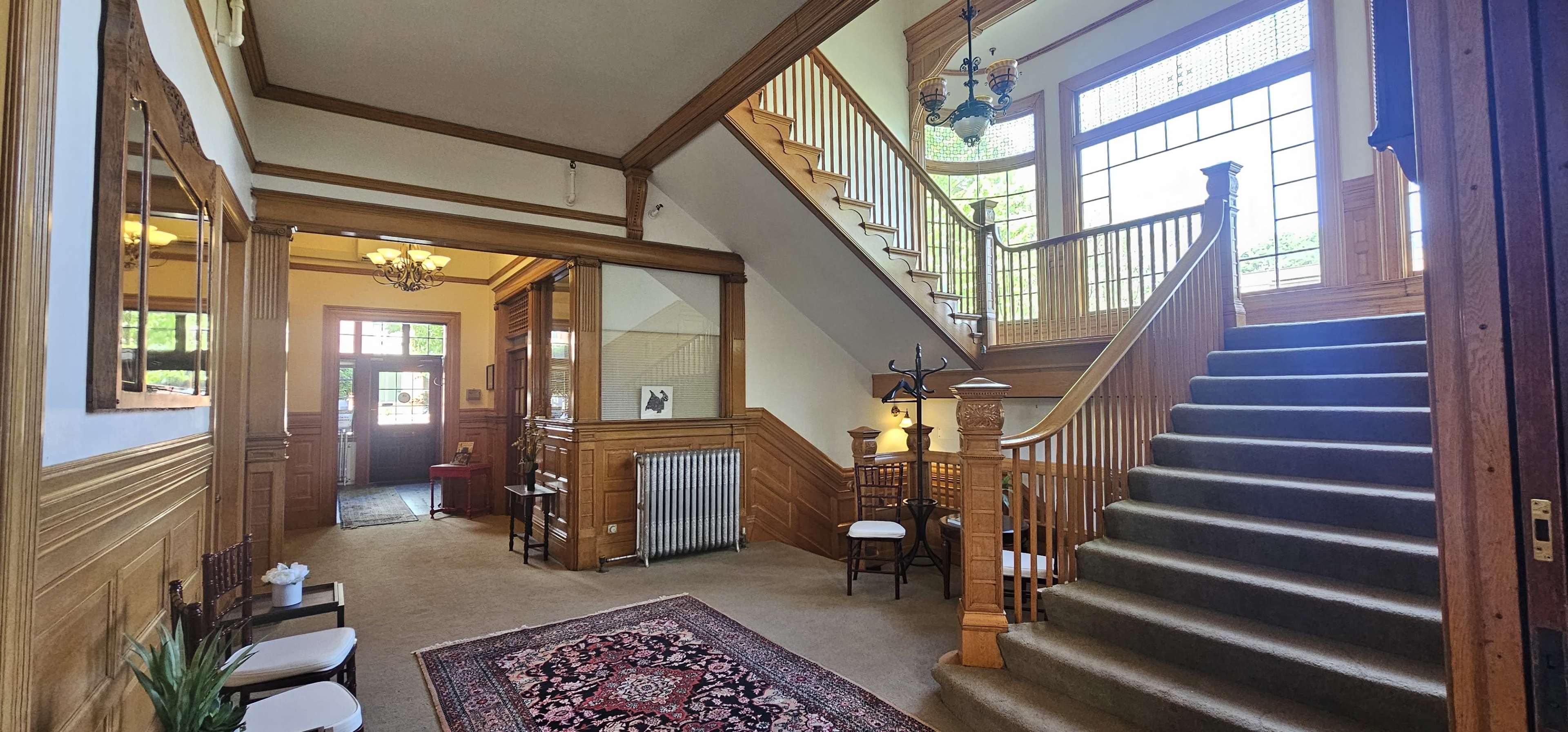 The image shows a spacious entrance hall with a grand staircase, wooden paneling, and large windows allowing natural light to illuminate the area.