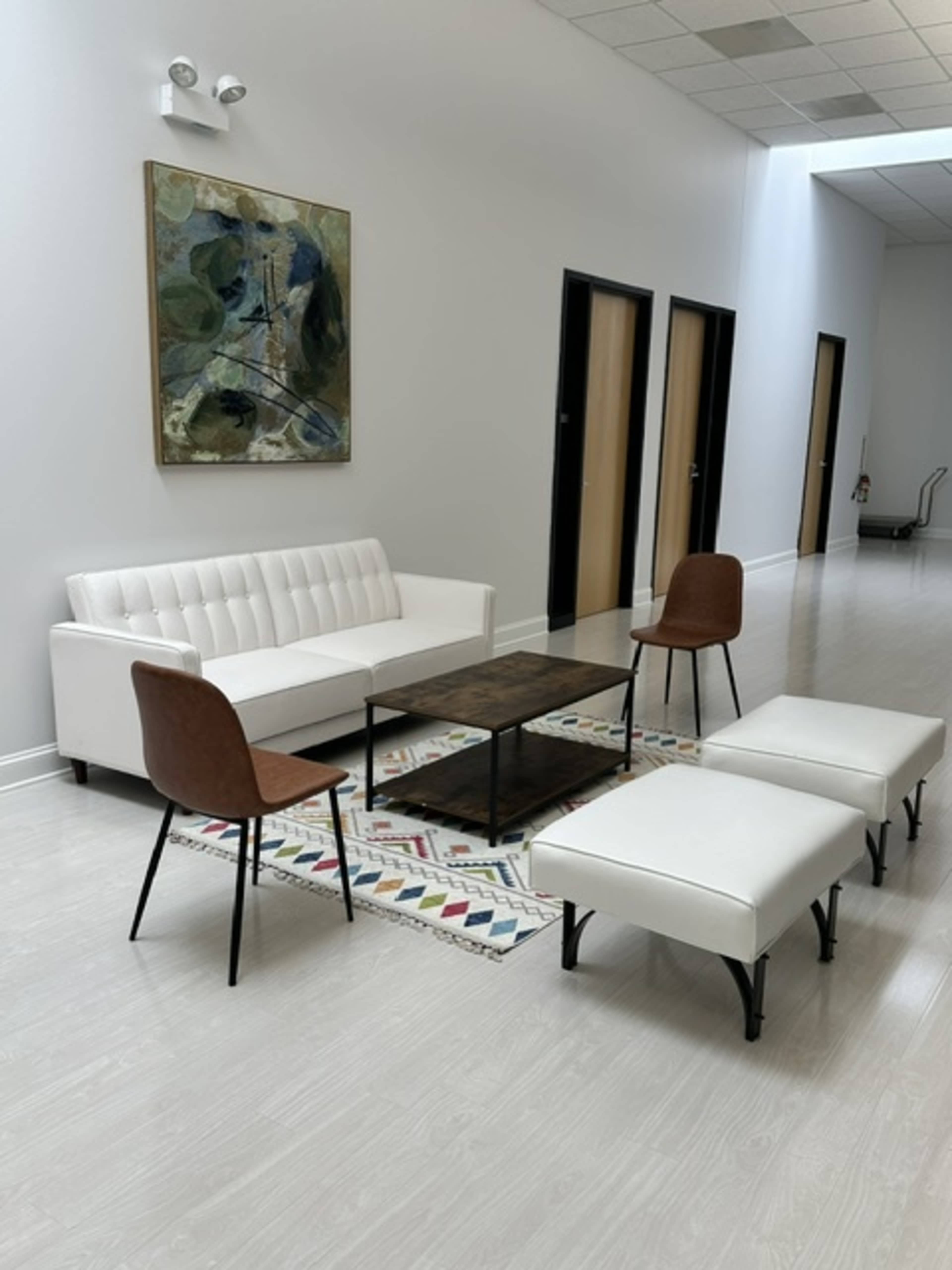 The image shows a modern seating area with a white sofa, two brown chairs, a wooden coffee table, and two white ottomans arranged on a decorative rug in a well-lit hallway.