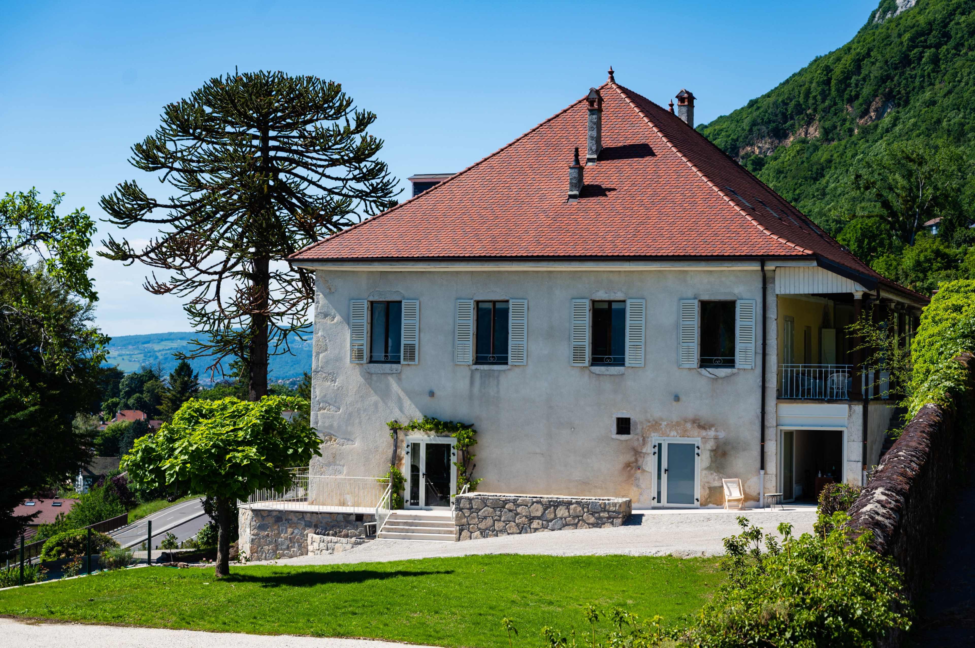 A large two-story house with a red-tiled roof and white walls is surrounded by greenery and a stone pathway, with a prominent tree nearby.