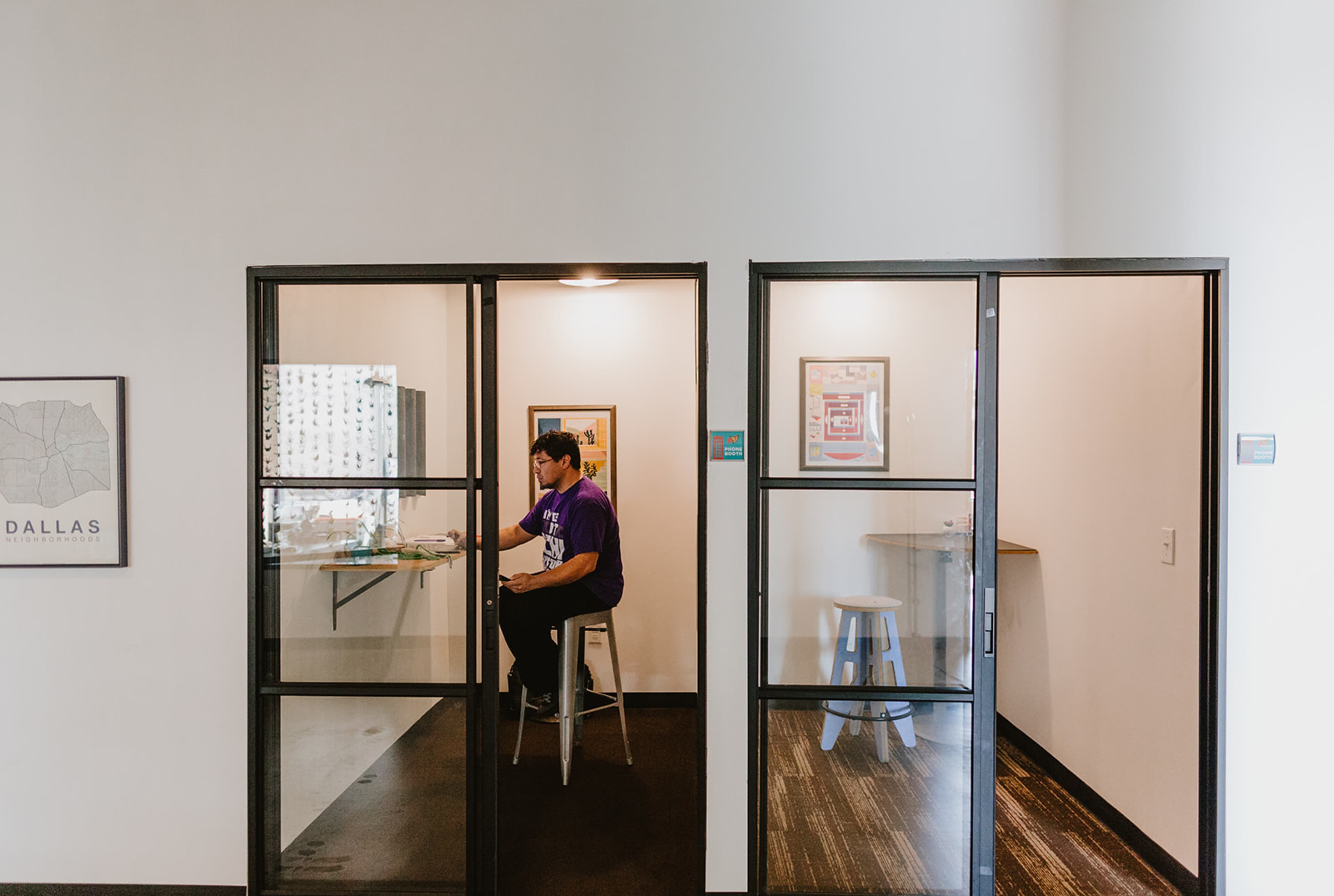 A person sits at a desk inside a glass-walled office space while another room, featuring a small circular table and stool, is visible adjacent to it.
