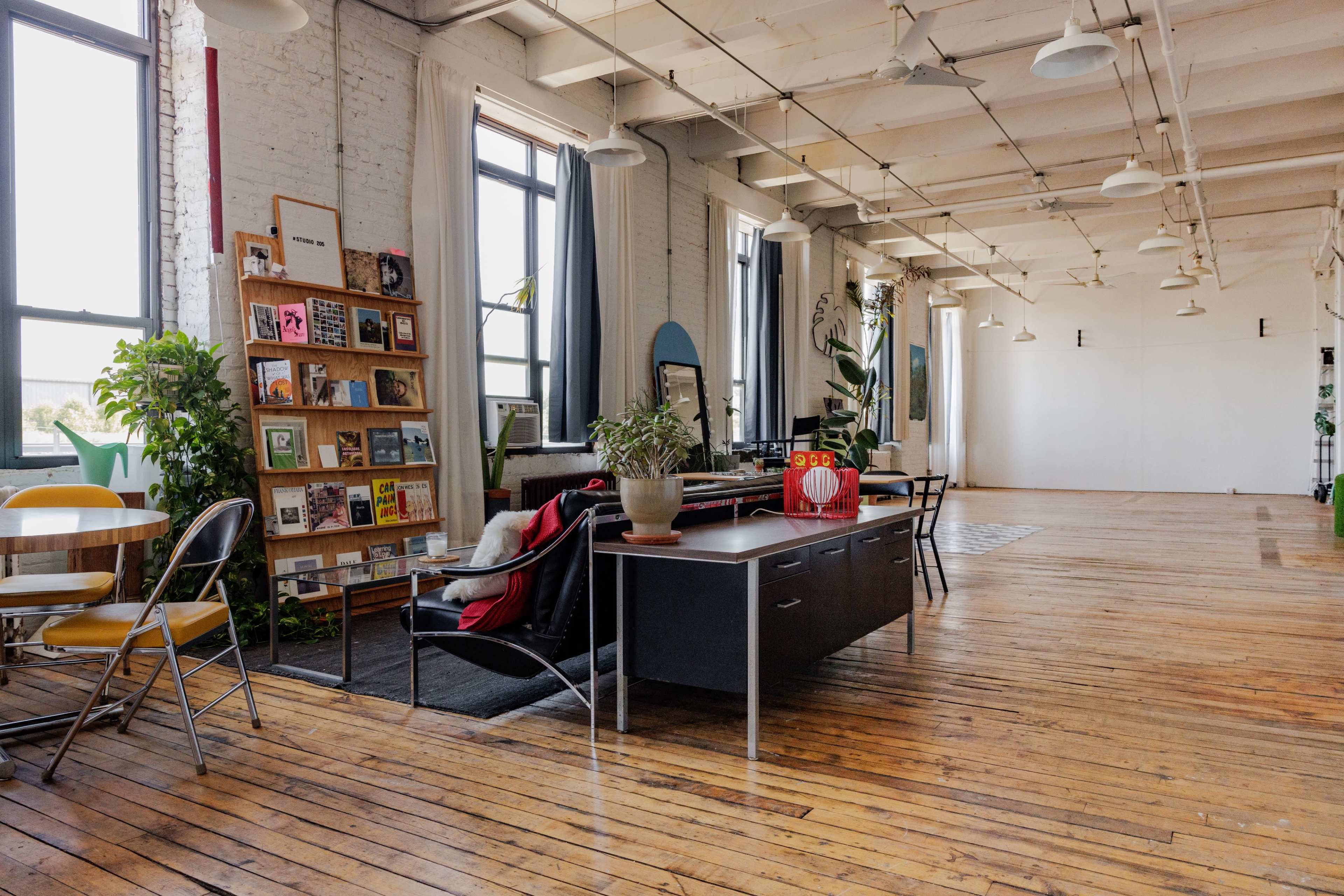 A spacious room with large windows, wooden floors, a bookshelf filled with magazines, and a desk with a chair and plants.