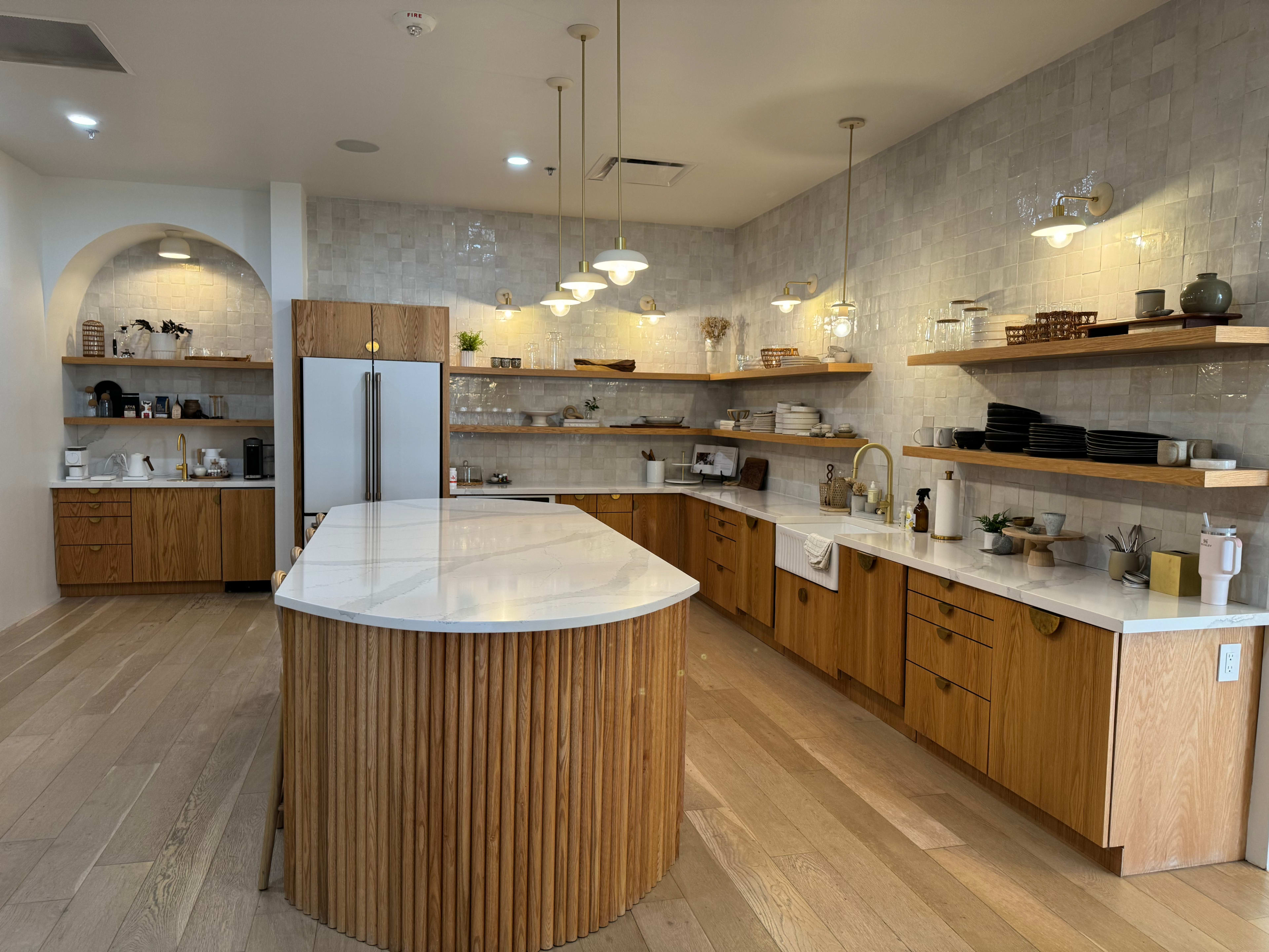 A modern kitchen features a large island with a white countertop, wooden cabinetry, open shelving with dishware, and multiple light fixtures.