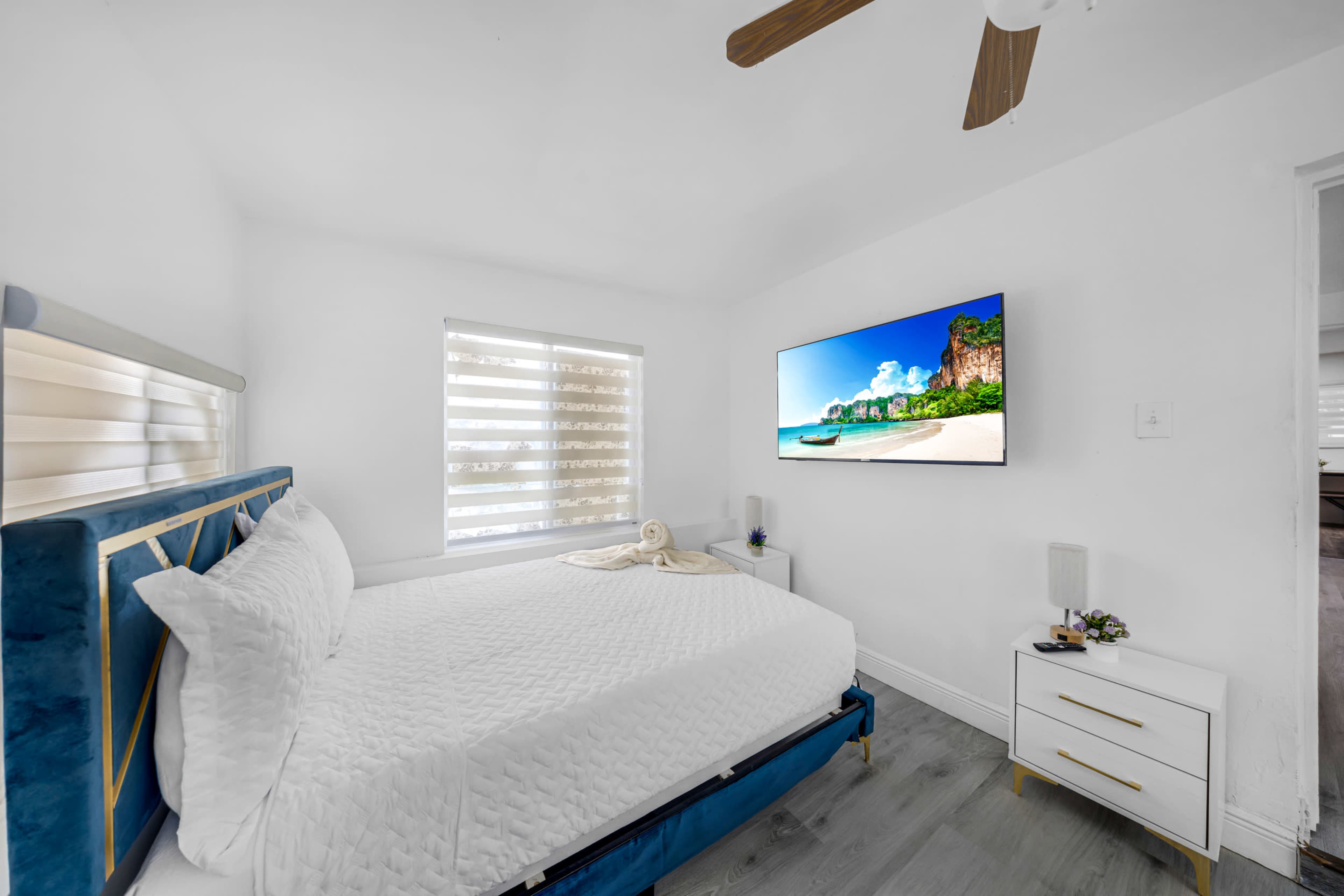 The bedroom features a neatly made bed with a blue headboard, a wall-mounted TV, and a white bedside table in a bright, minimalistic space.