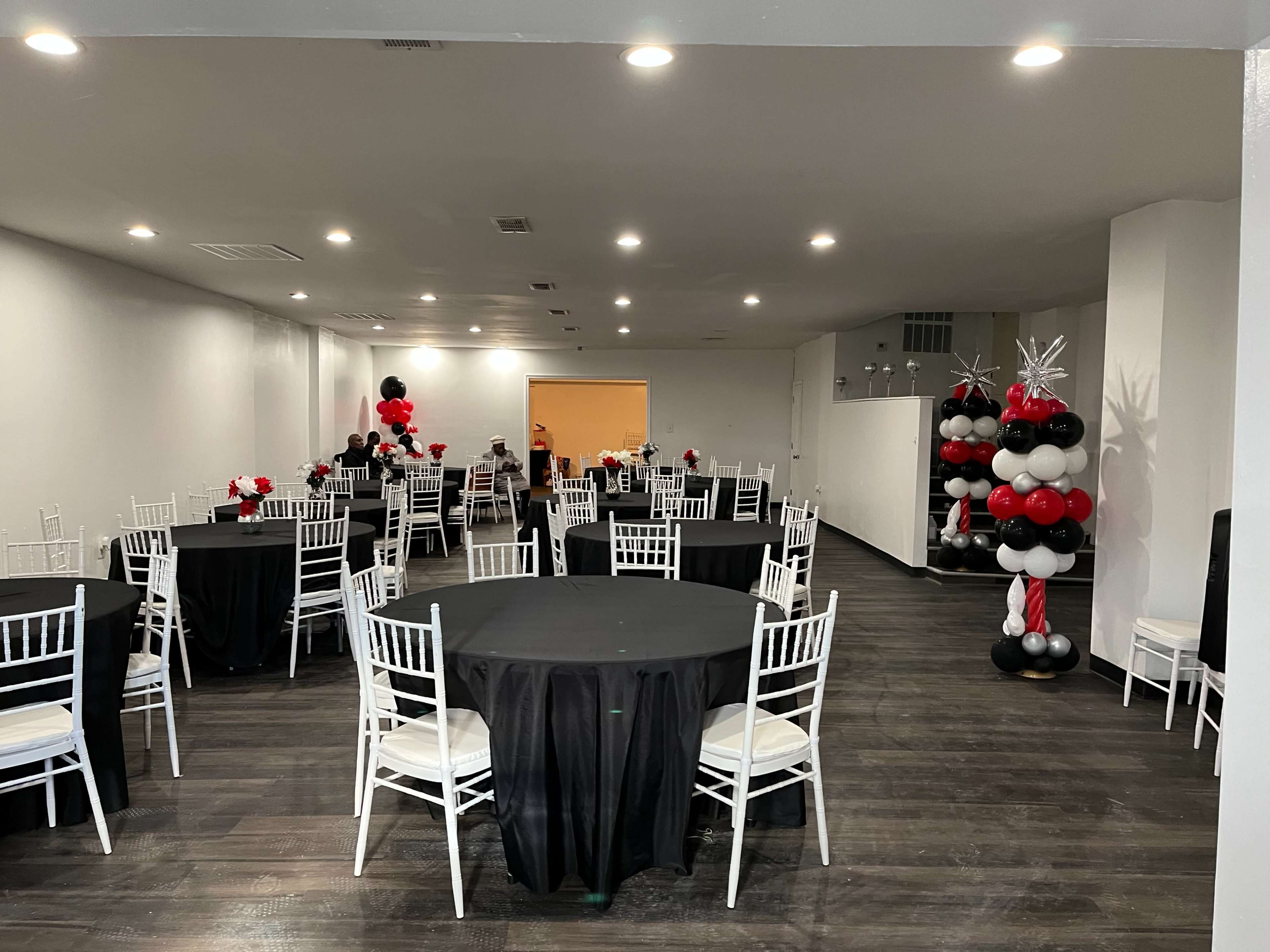 A spacious event room features black round tables with white chairs, decorated with red and black balloons arranged in columns.