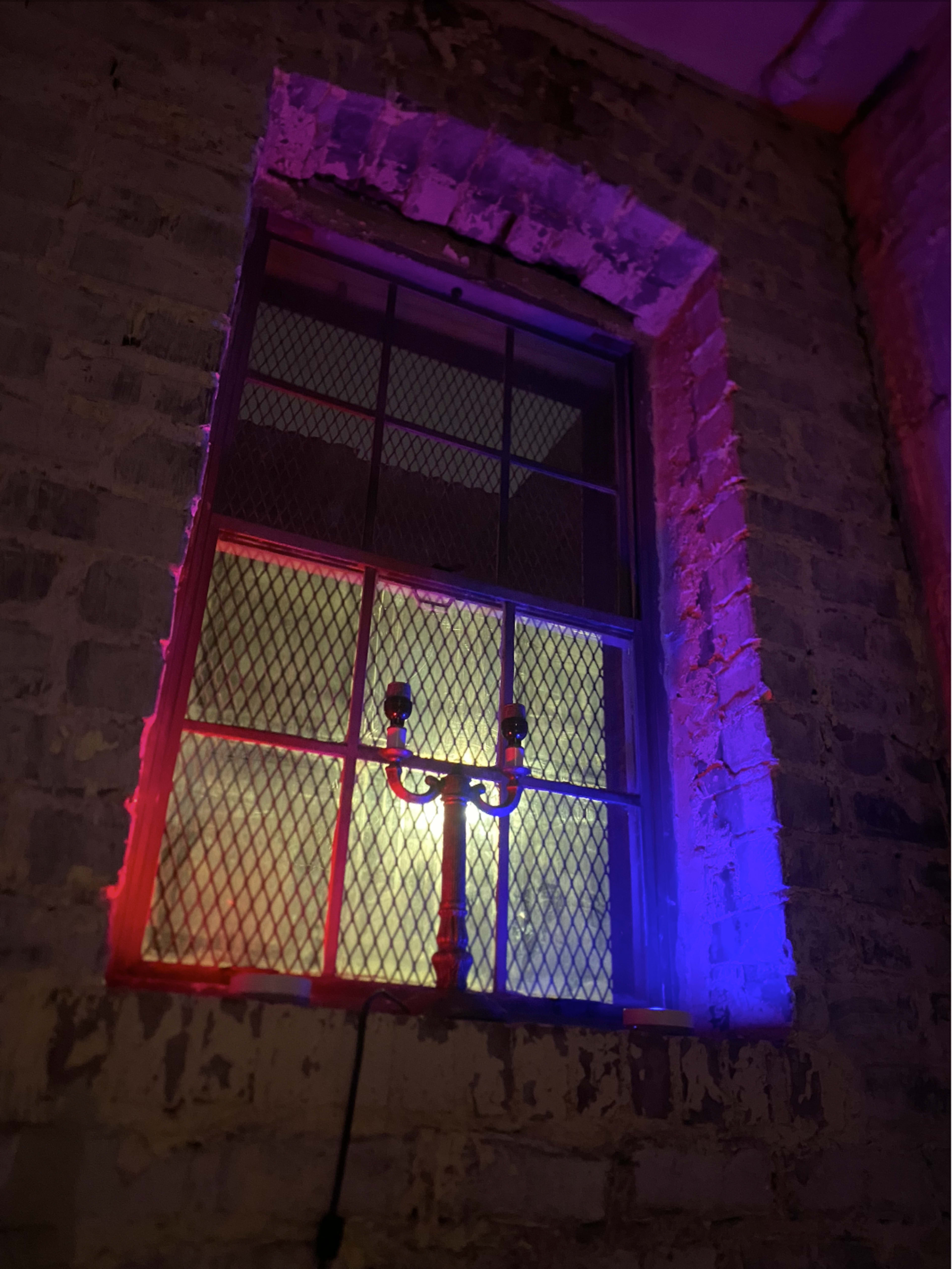 A window with a metal grid, illuminated by red and blue lights, set against a textured brick wall.
