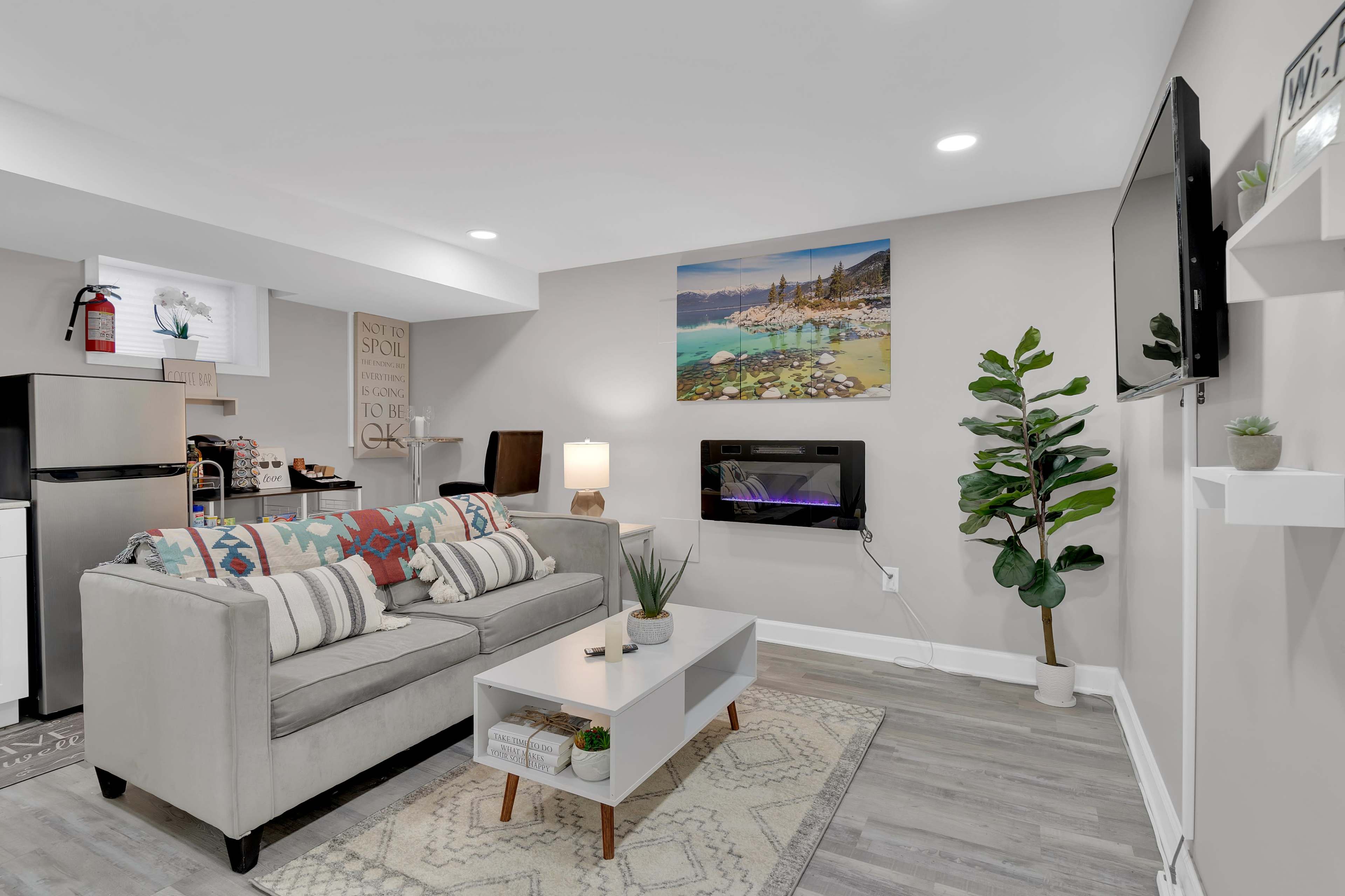 A modern living room with a light gray sofa, a coffee table, a wall-mounted TV, and a decorative plant, complemented by a painting of a coastal scene.