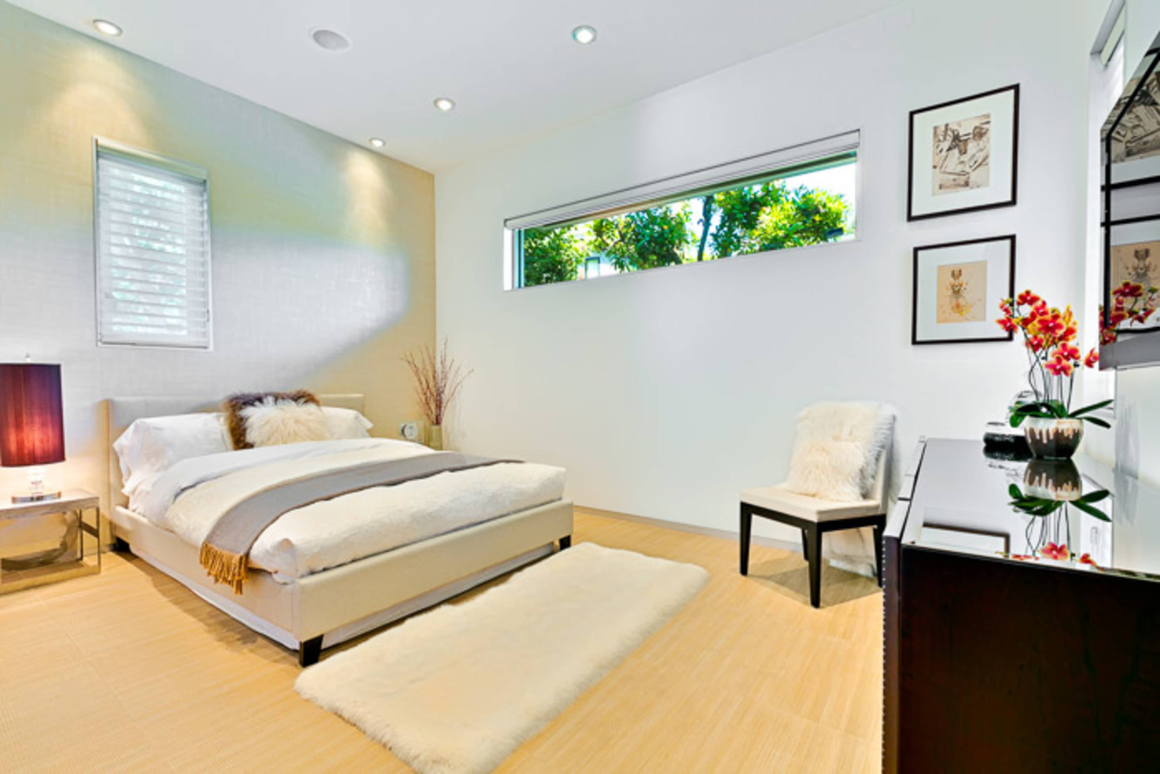 The image shows a modern bedroom with a bed, a chair, and a window bringing in natural light.