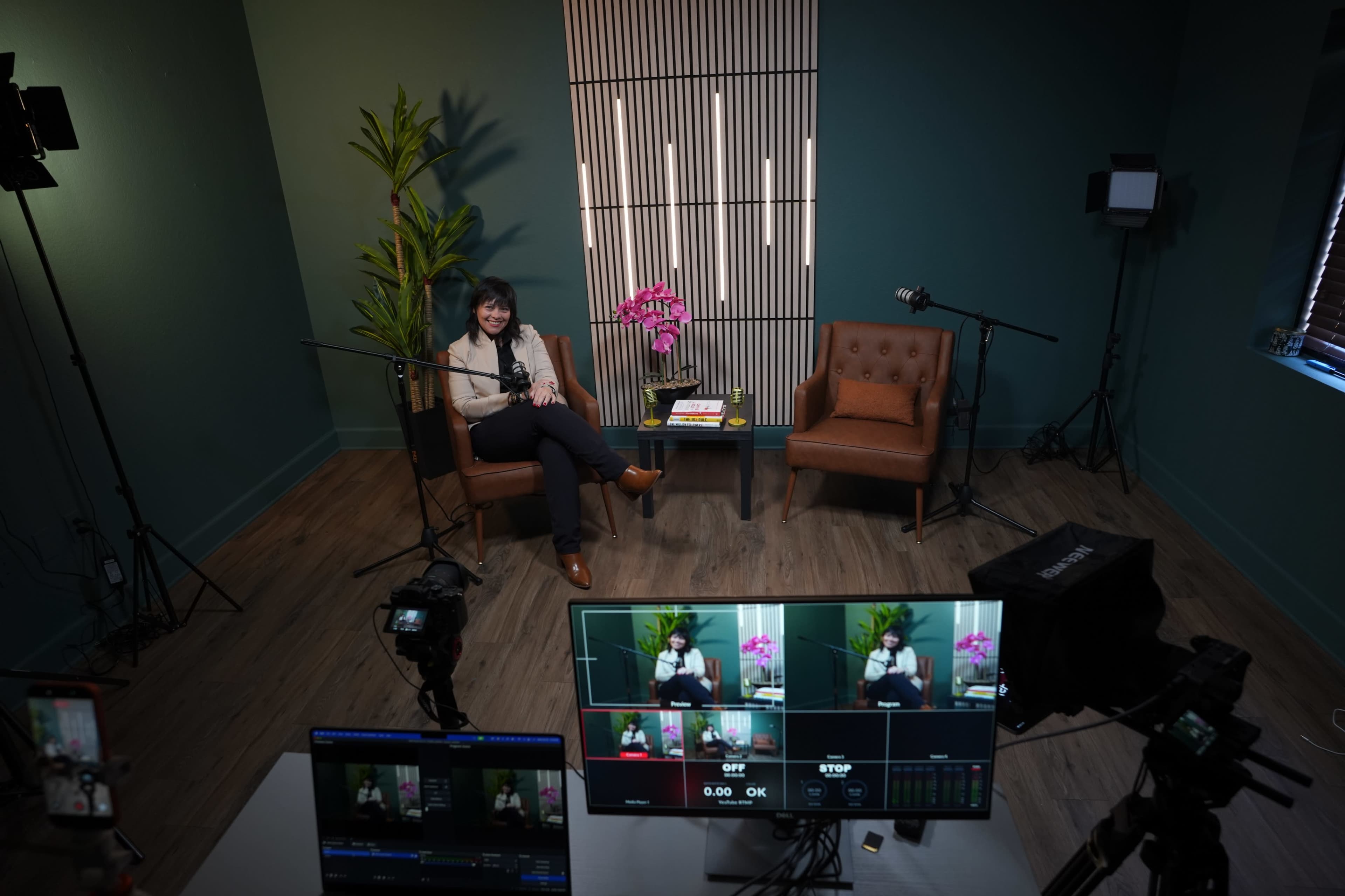 A woman sits in a studio setting with two chairs, a table, and several cameras and lights arranged for filming.
