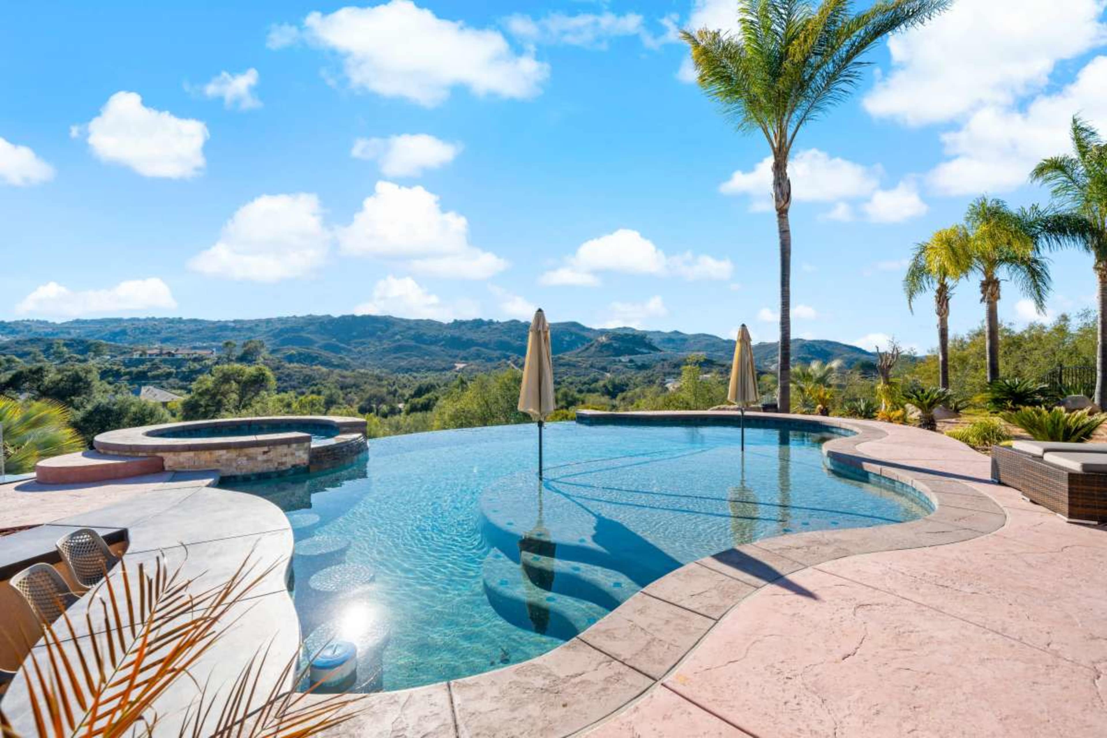 The image shows a modern swimming pool with a scenic backdrop of hills and palm trees under a blue sky.