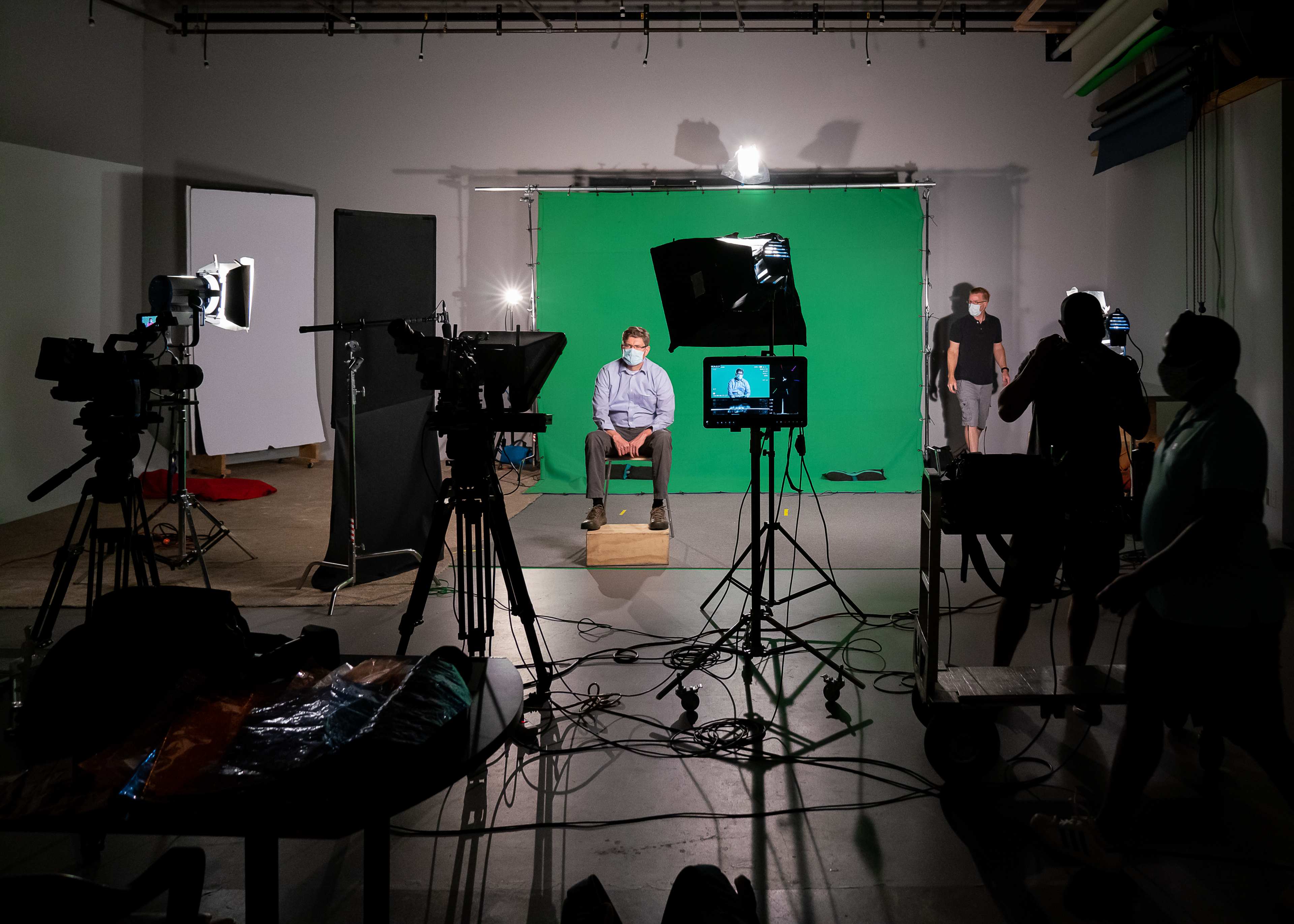 A person sits on a wooden box in front of a green screen while multiple camera setups and crew members are positioned nearby.