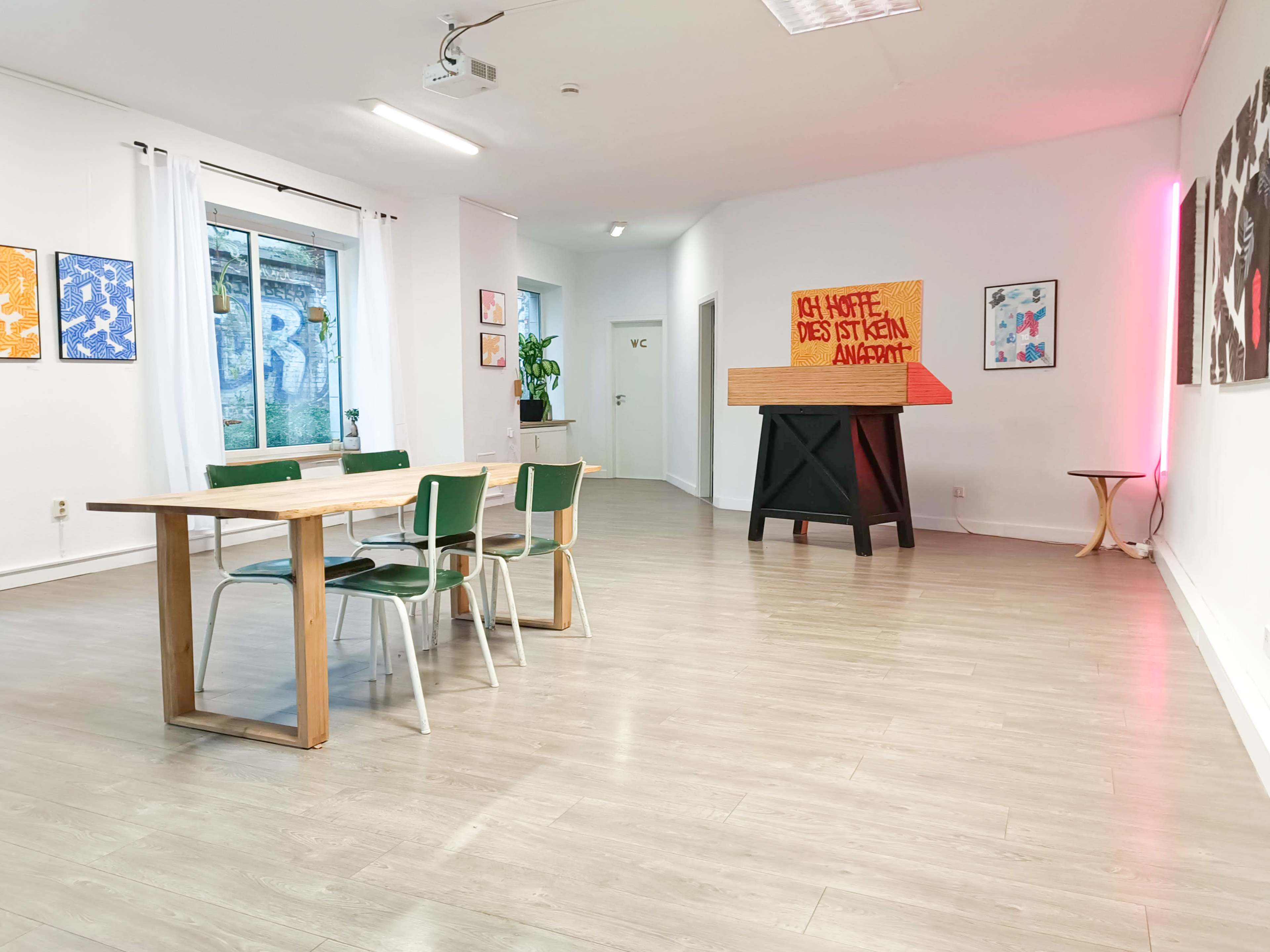 The image shows a modern, minimalist interior space with a wooden table surrounded by chairs, and a large, colorful artwork placed on an elevated stand against a white wall.