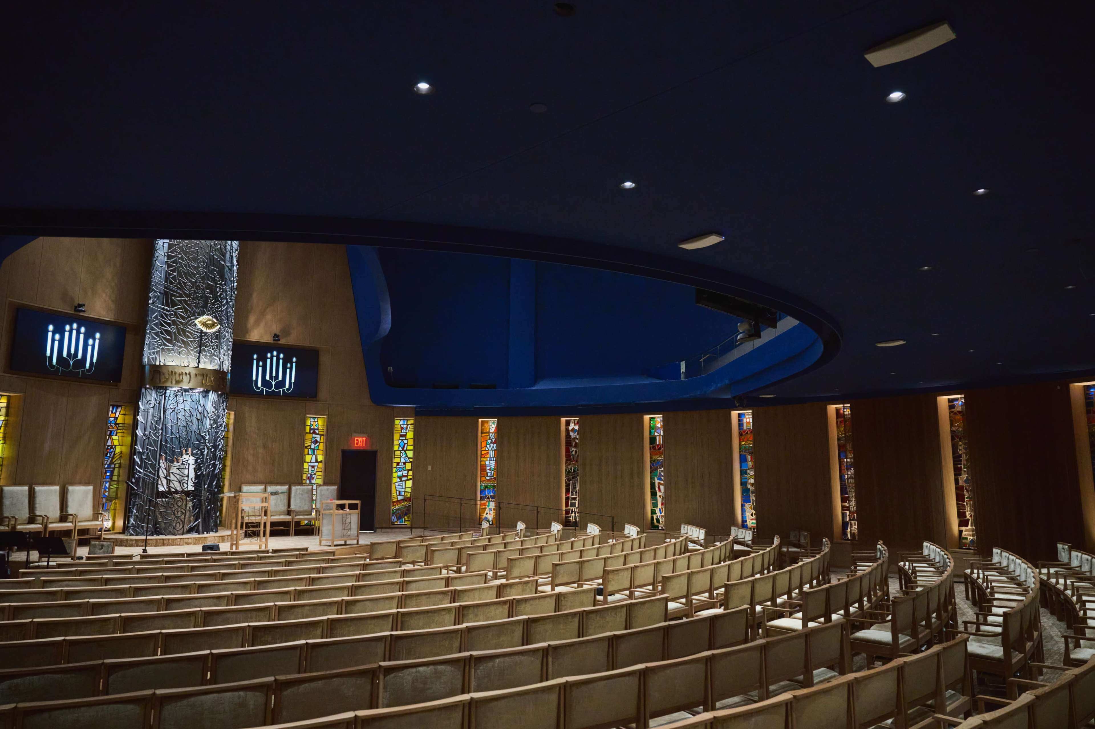 A modern interior of a synagogue with rows of wooden chairs, stained glass windows, and a central stage area.