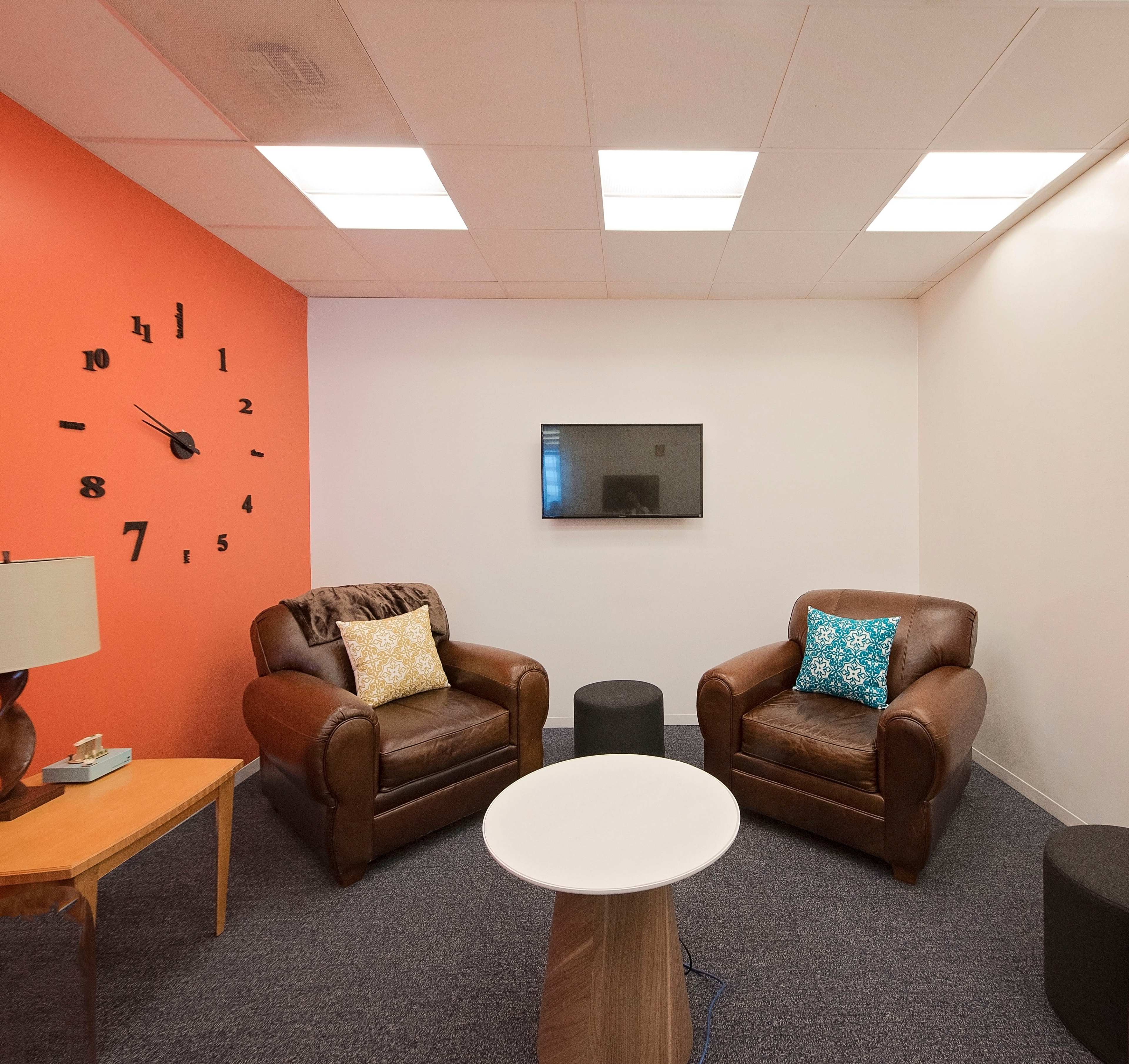 The image shows a small indoor lounge area featuring two brown leather chairs, a round white table, a wall clock, and a flat-screen television on the white wall.