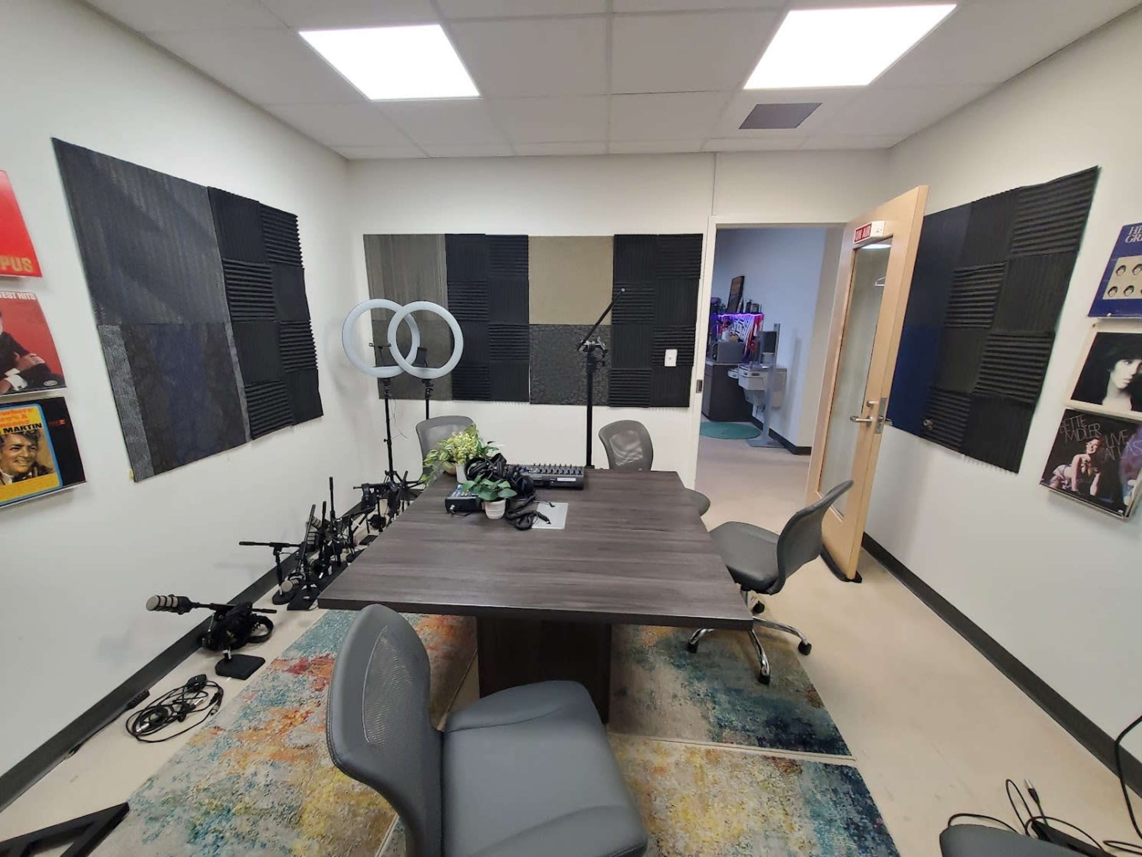 A small recording studio with a large table, two chairs, soundproofing panels on the walls, and various audio equipment arranged around the room.