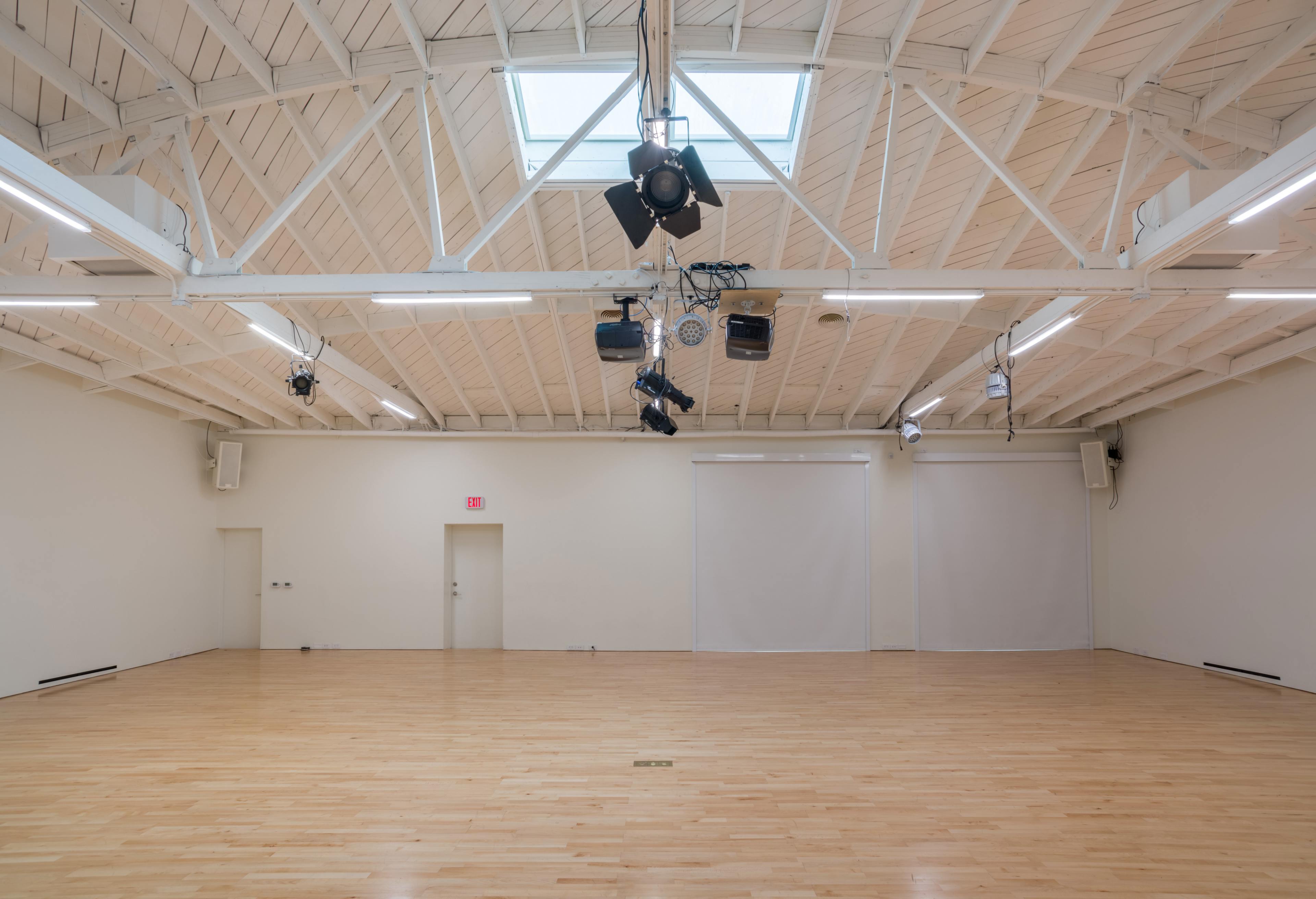 A spacious, empty room with wooden flooring, high ceiling beams, and overhead lights.