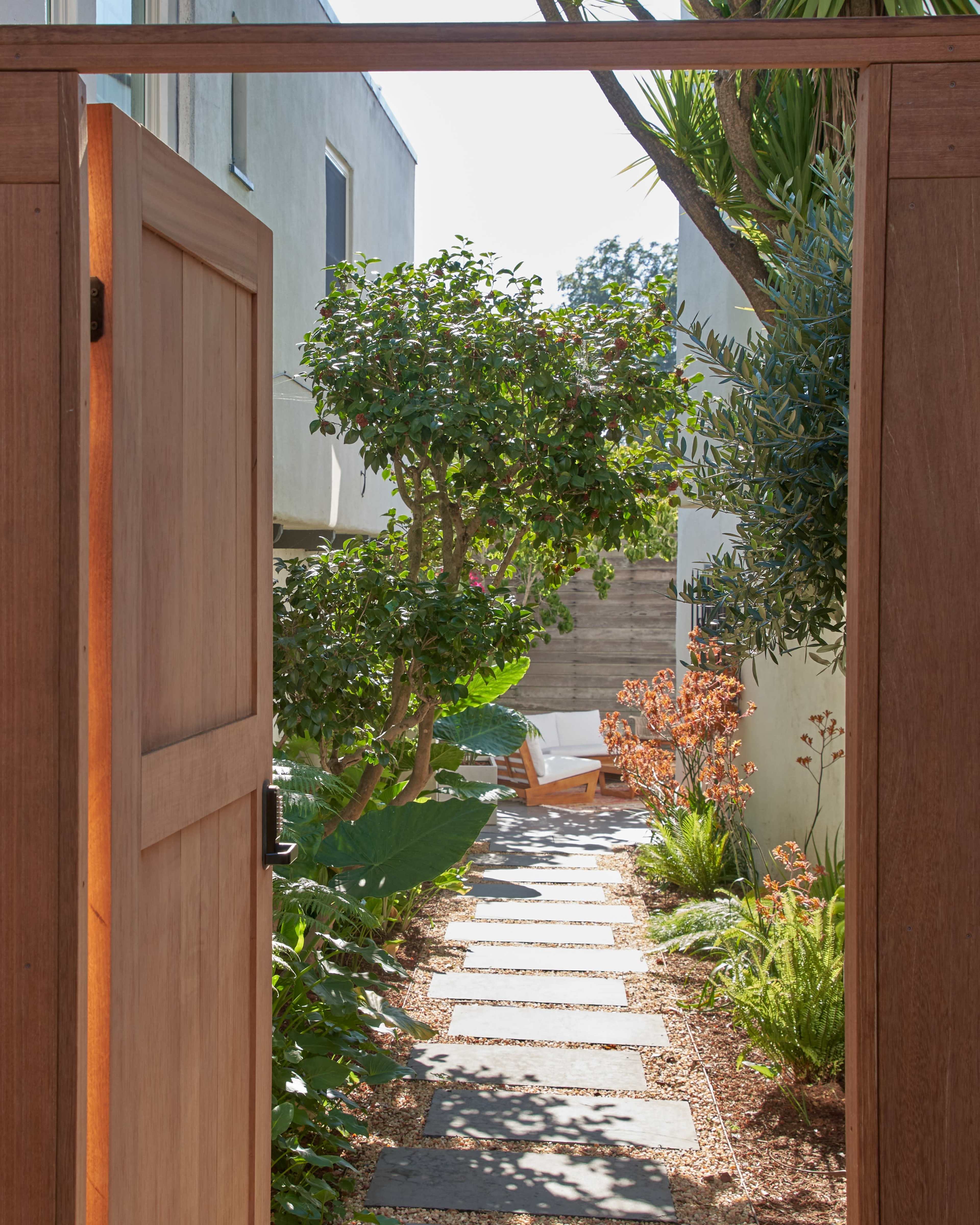A wooden gate opens to a stone pathway lined with plants leading to a chair under a tree.