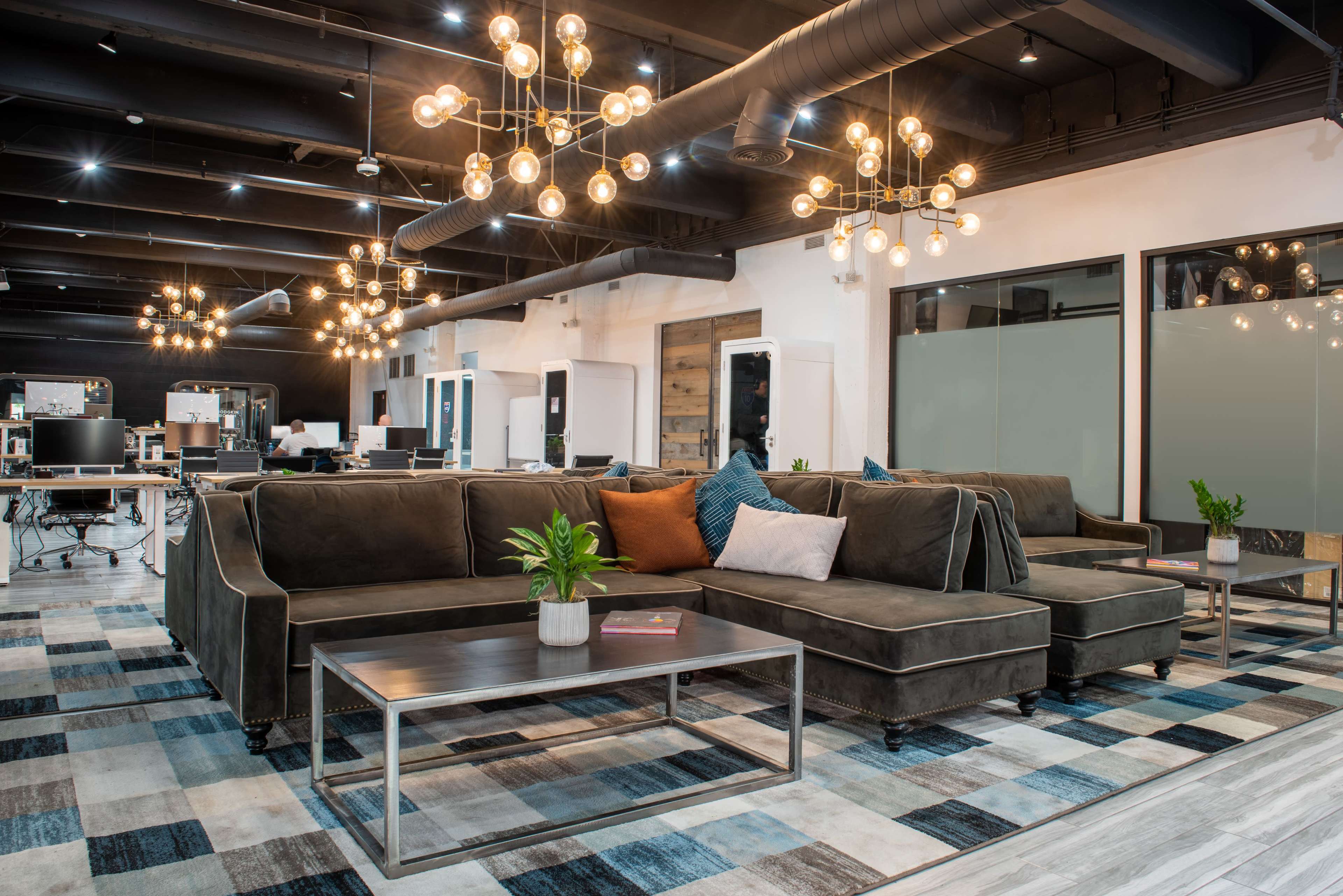 The image shows a modern office lounge area with a large sectional sofa, decorative lighting fixtures, and a coffee table on a patterned rug.