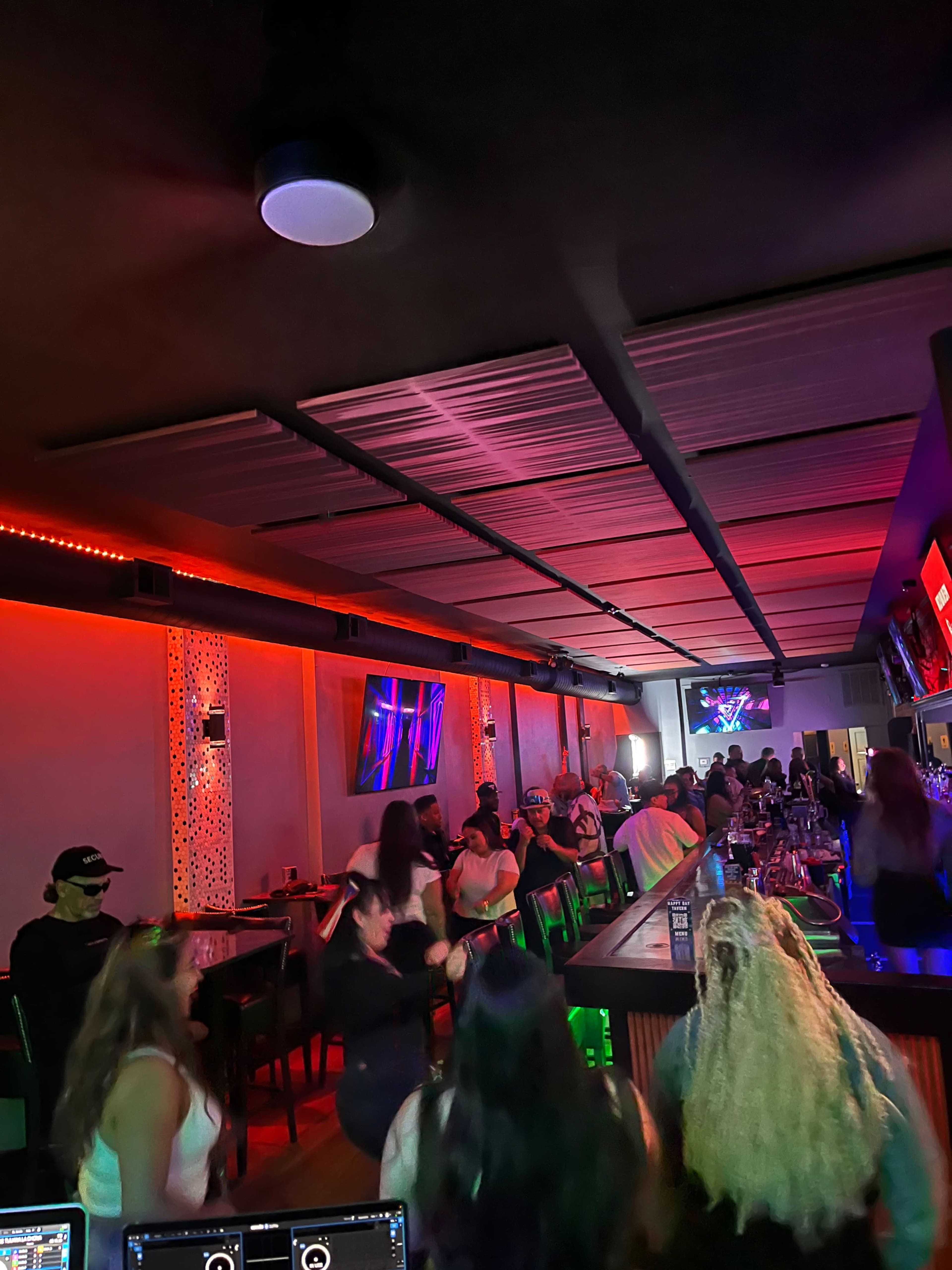 A crowded bar scene features patrons dancing under vibrant red and blue lighting, with a DJ setup visible in the foreground.