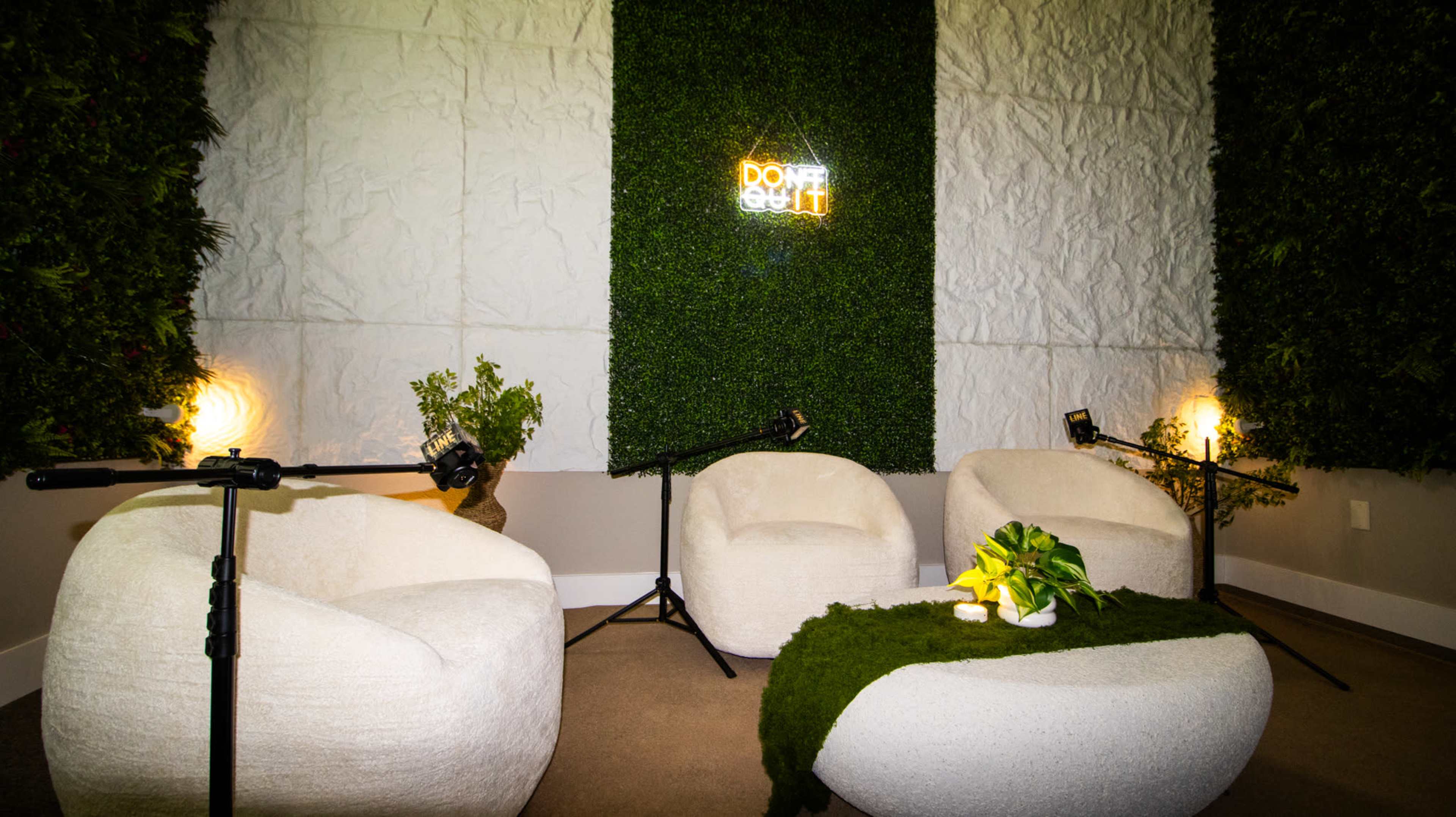 The image shows a cozy room featuring three white, rounded chairs arranged around a stone-like coffee table, with green plants on the table and a moss-covered wall in the background, illuminated by soft lighting.