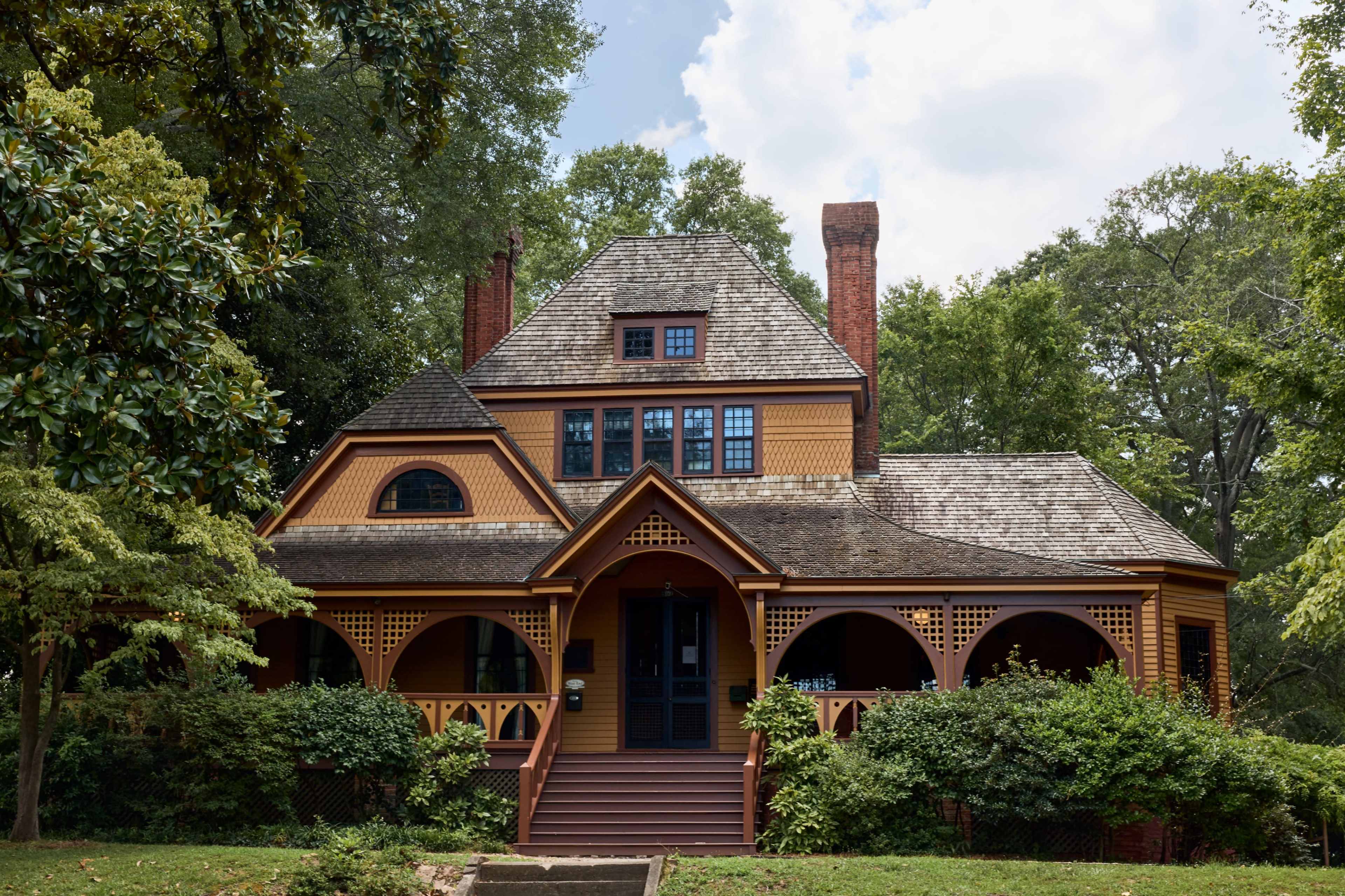 The image shows a large, Victorian-style house with a multi-gabled roof, vibrant yellow and brown exterior, and surrounded by greenery.