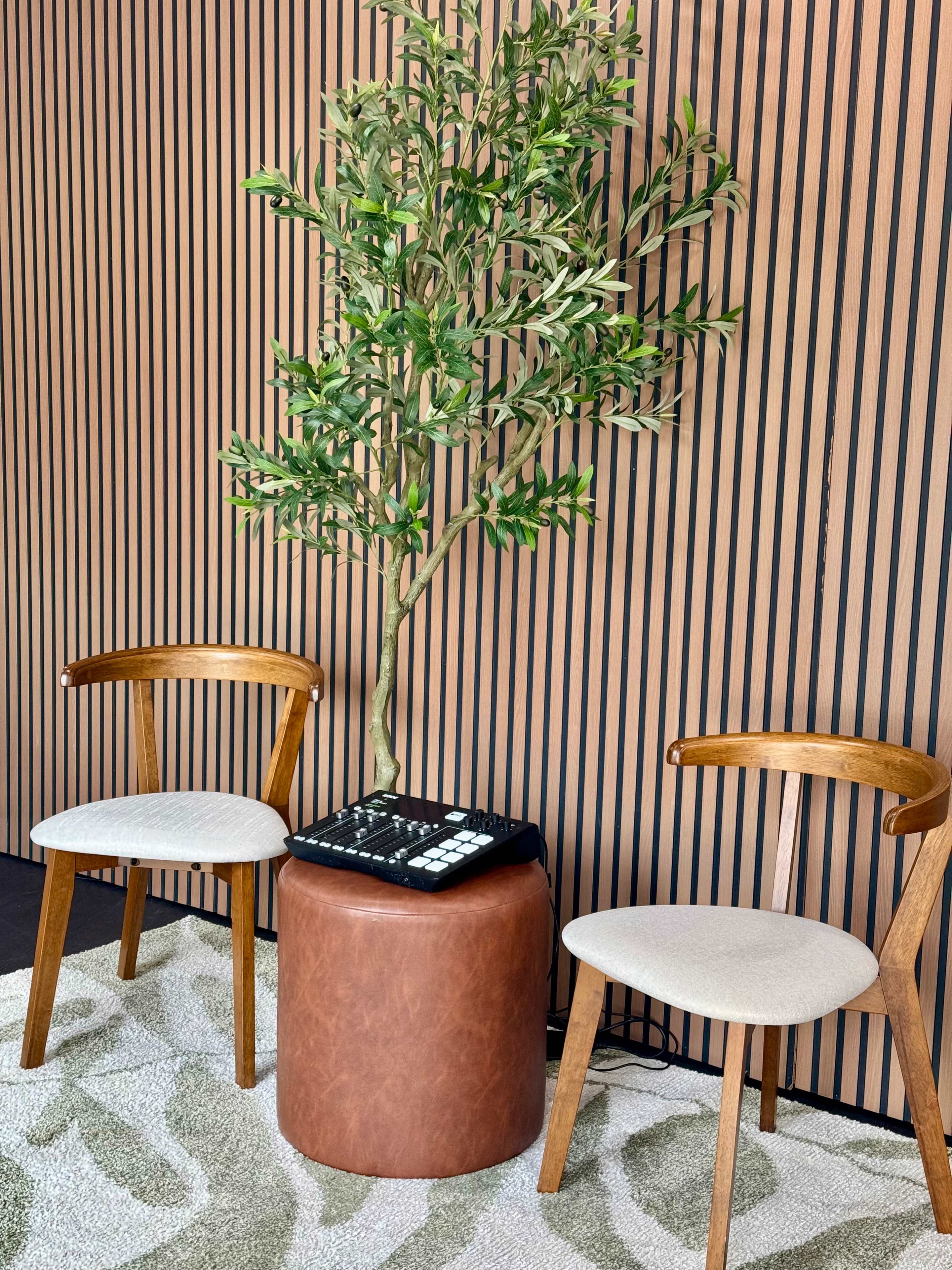 The image shows two wooden chairs and a round, leather ottoman placed beside a potted artificial tree against a backdrop of vertical striped wood paneling.
