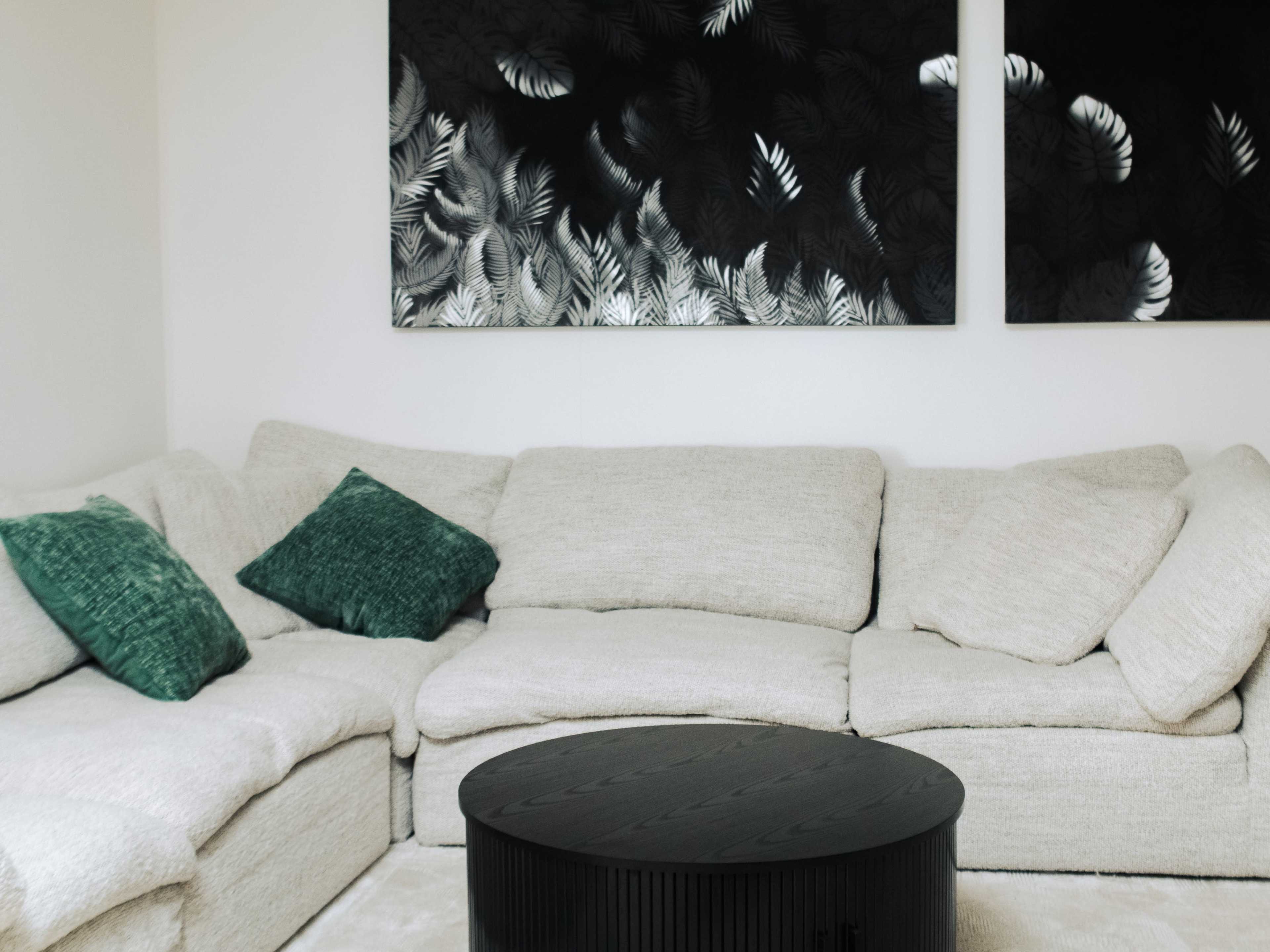 The image shows a cozy living room with a light-colored sectional sofa adorned with green cushions, facing a round black coffee table and two abstract leaf-patterned artworks on the wall.