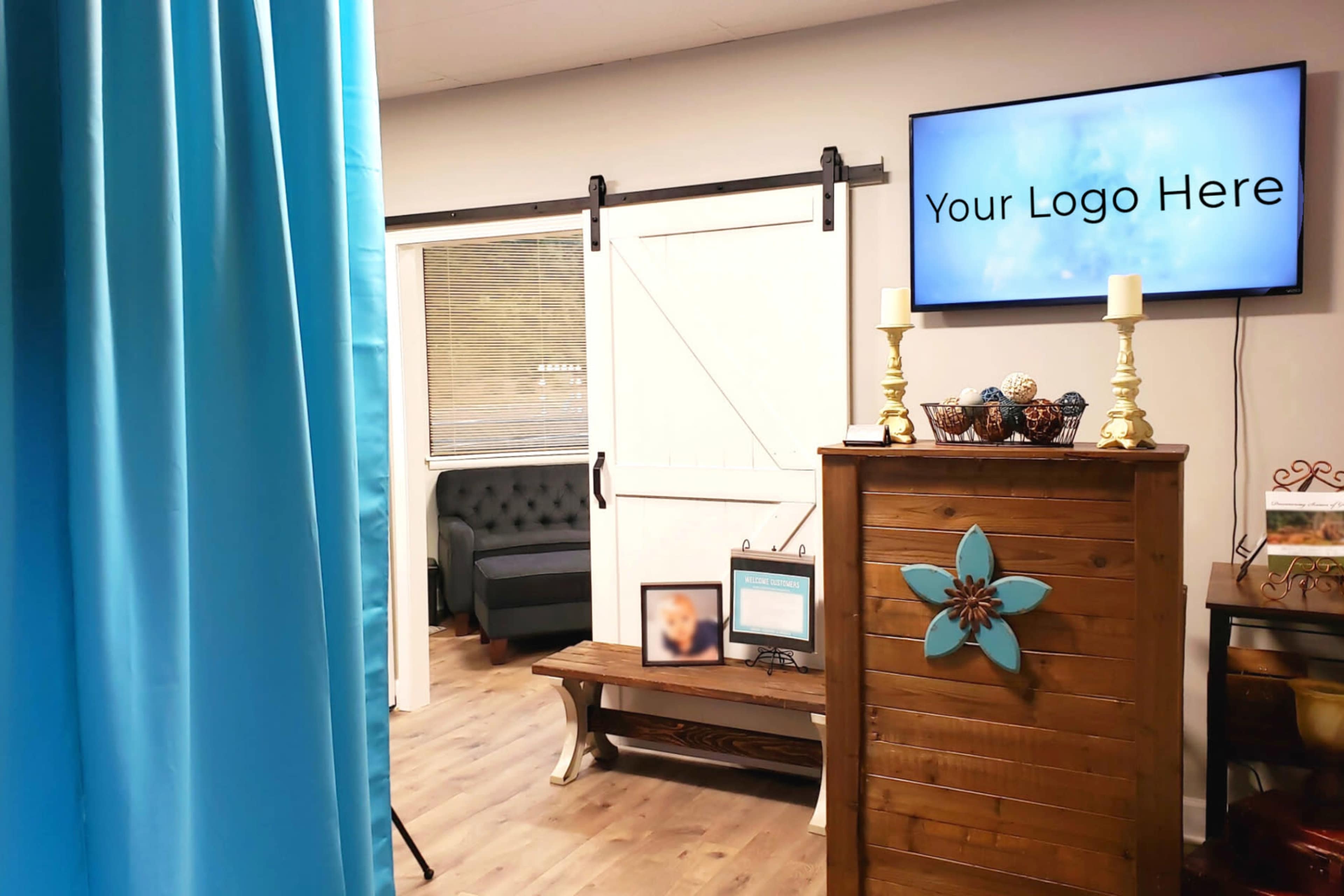 A waiting area with a comfortable couch, a decorative wooden cabinet featuring a blue flower design, and a television mounted on the wall displaying "Your Logo Here."