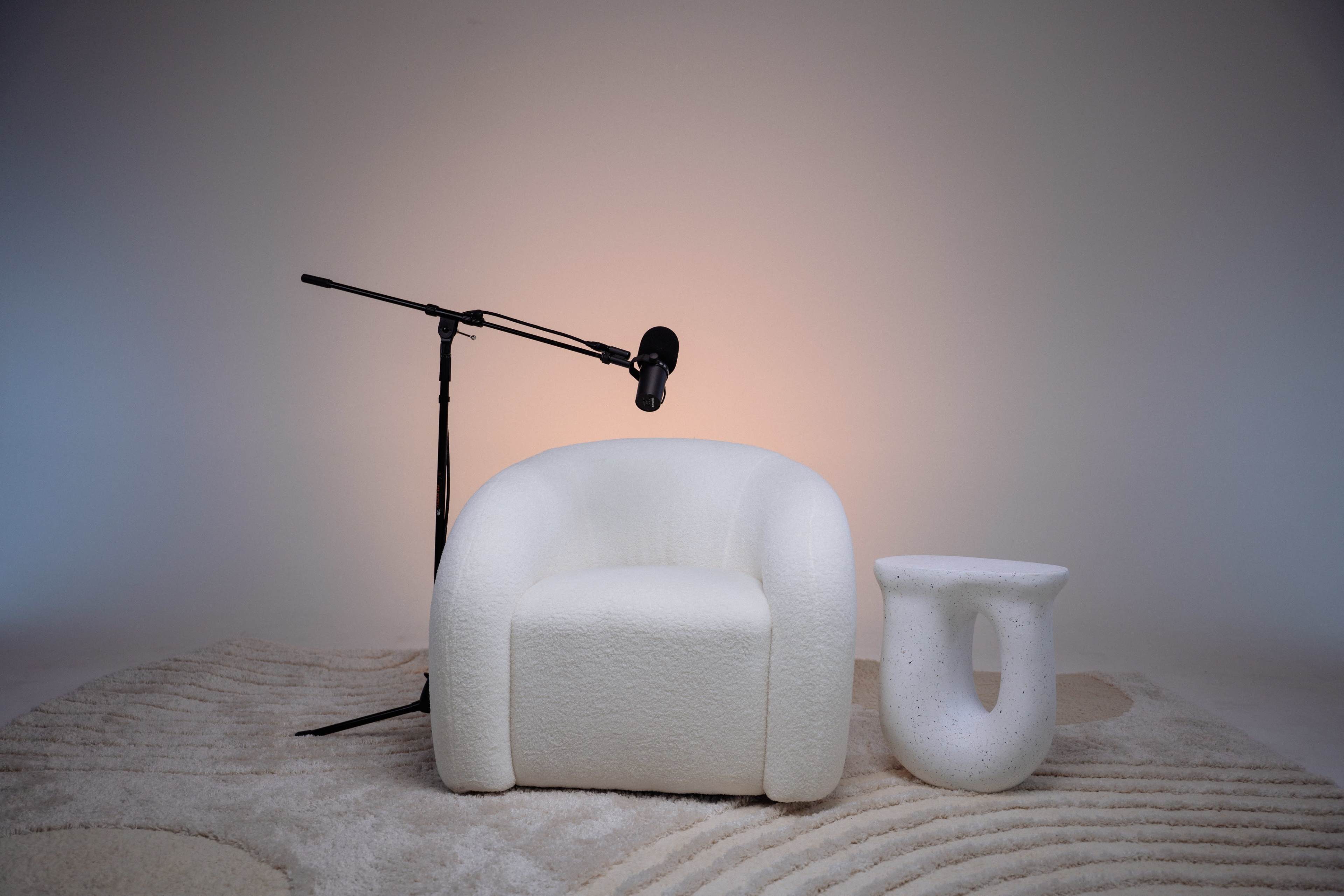The image features a white, rounded armchair next to a small, white side table, accompanied by a microphone on a stand against a softly lit background.