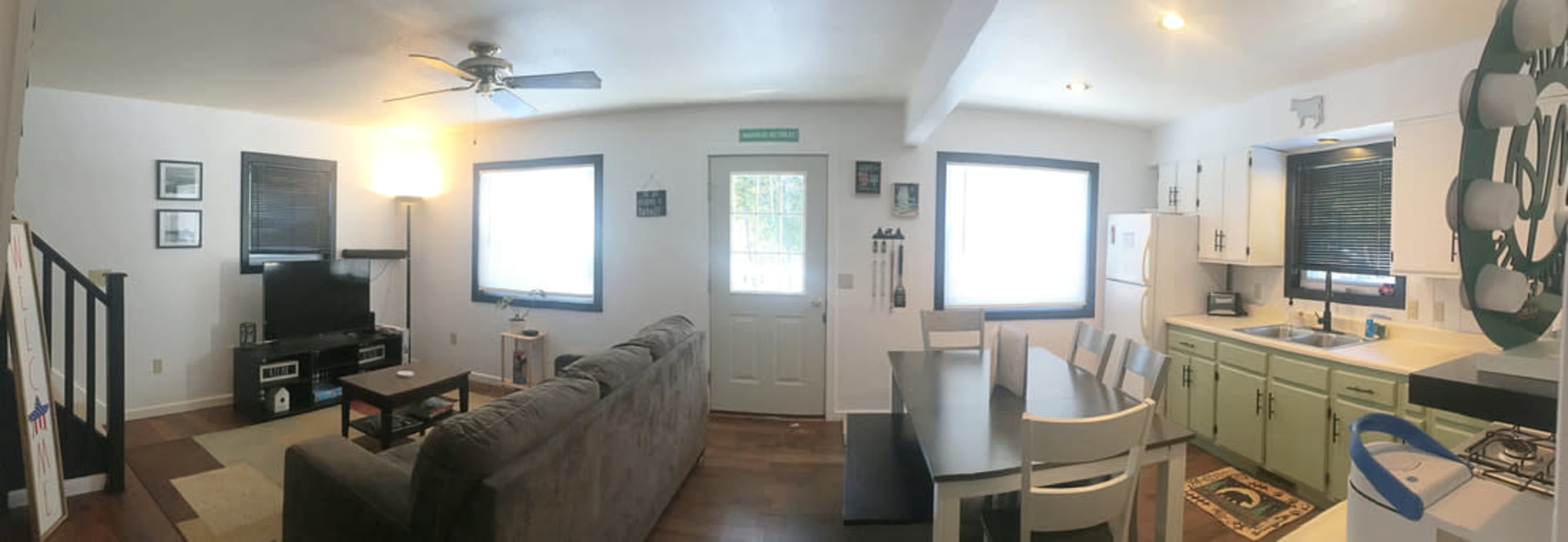 A panoramic view of a living area and kitchen with a sofa, television, dining table, and appliances in a well-lit space.