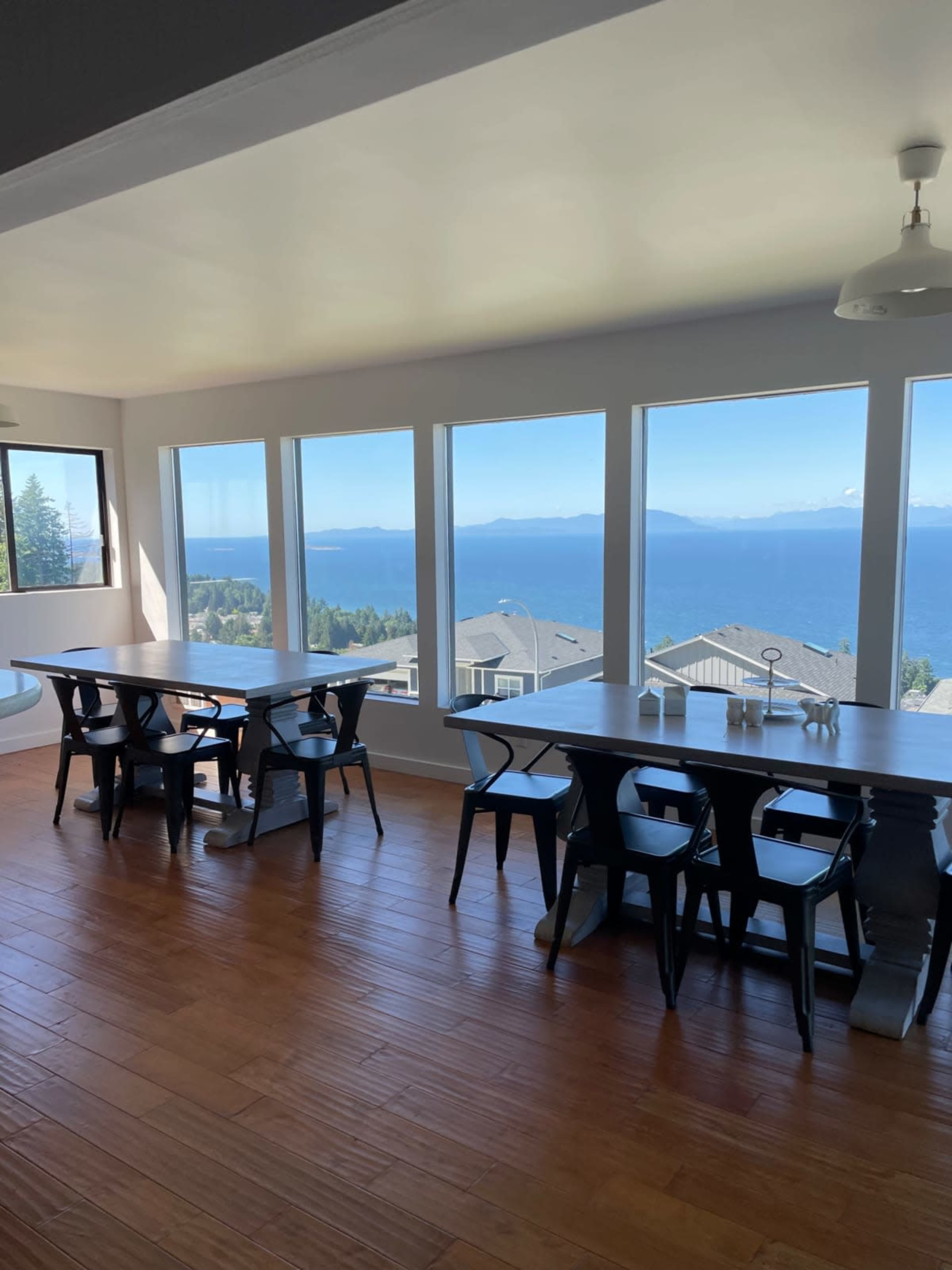 A bright, spacious room features two long wooden tables and large windows offering a view of a serene lake and distant mountains.