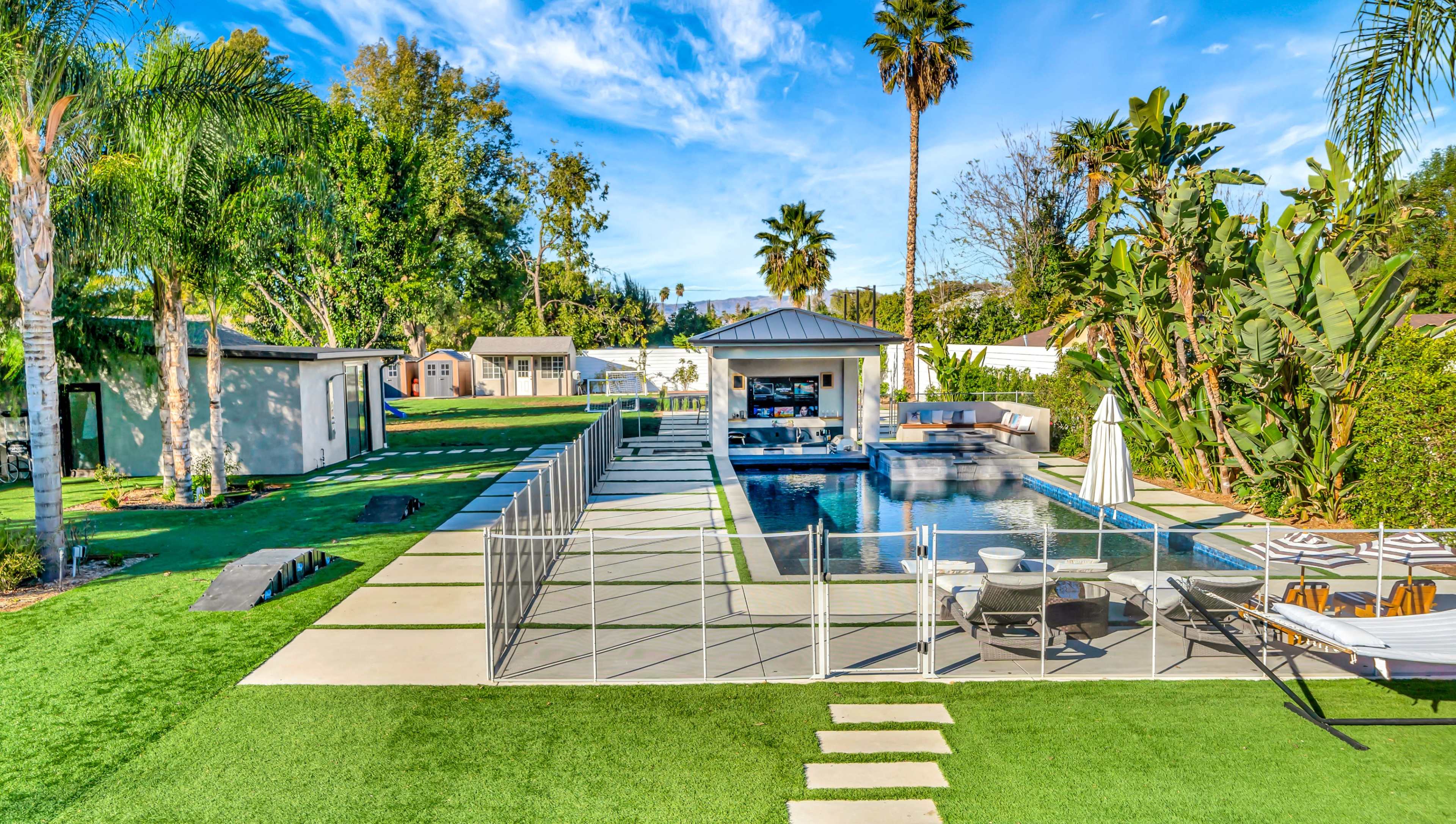 A modern backyard with a swimming pool, surrounded by palm trees, green grass, and a poolside area featuring lounge chairs and a gazebo.