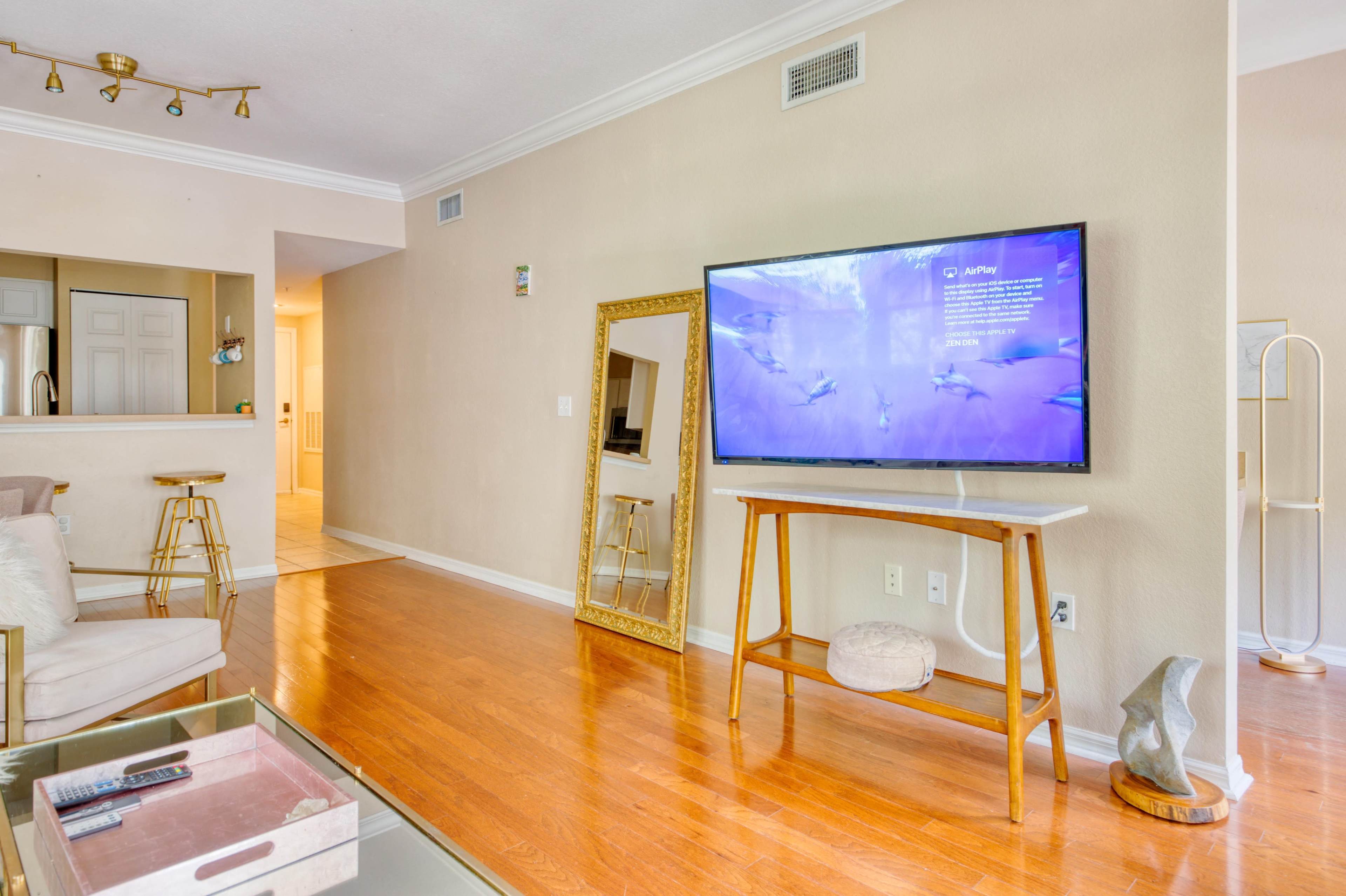 A living room with a large TV mounted on a stand, a mirrored wall decor, and light wooden flooring.