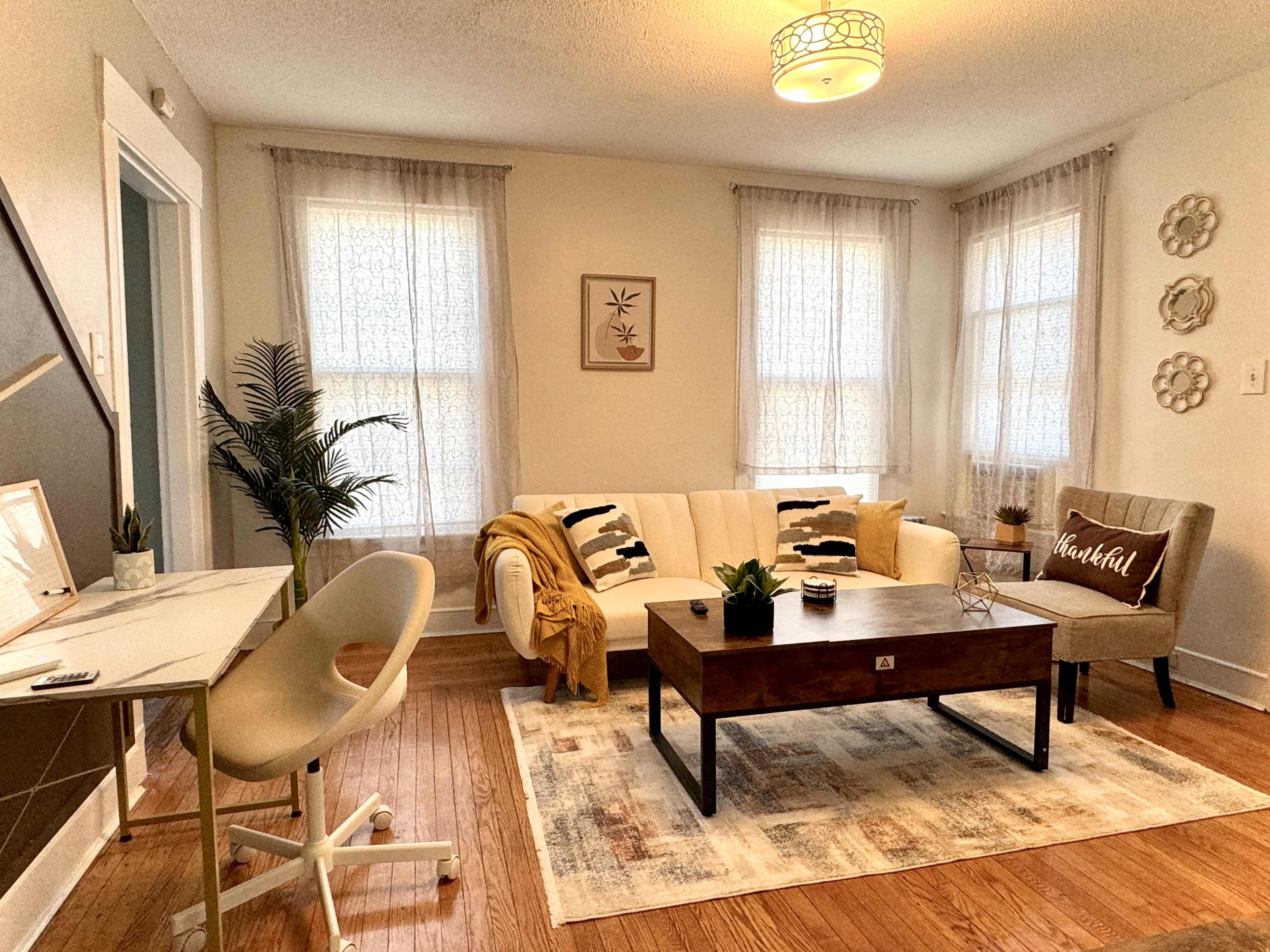 A cozy living room featuring a white sofa with black and white pillows, a wooden coffee table, a small armchair, and a working desk, all arranged around a patterned area rug.