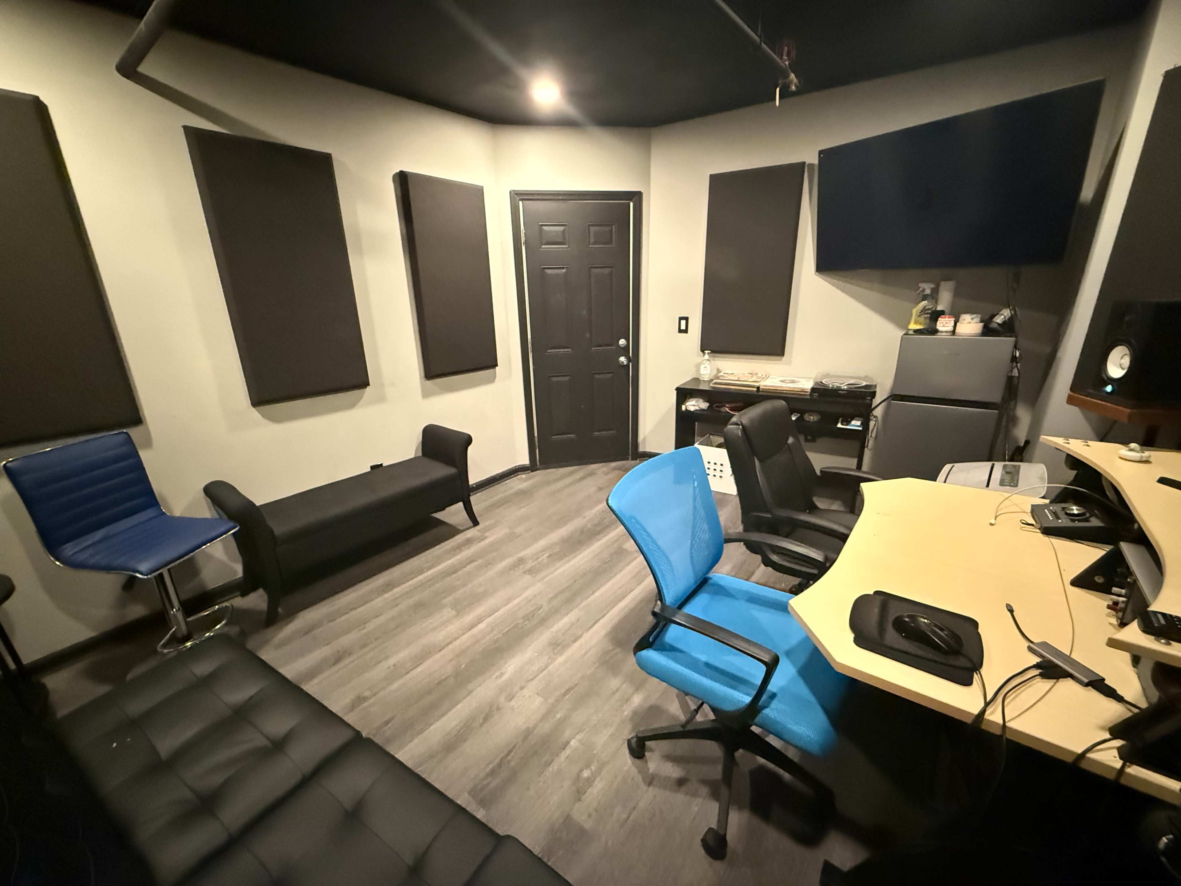 The image shows a recording studio featuring a control desk, several chairs, soundproofing panels on the walls, and a closed door.
