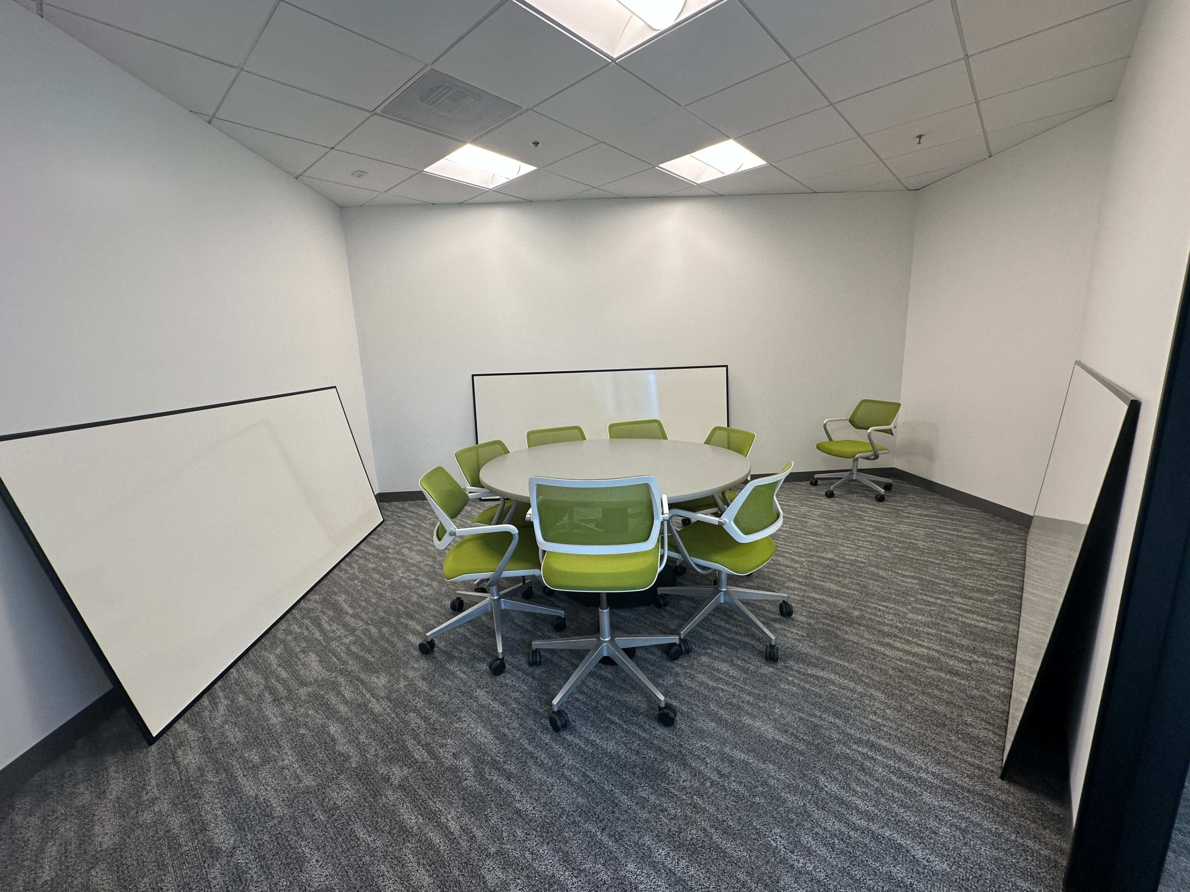 A modern meeting room features a circular table surrounded by green and white chairs, with two large white boards leaning against the walls.