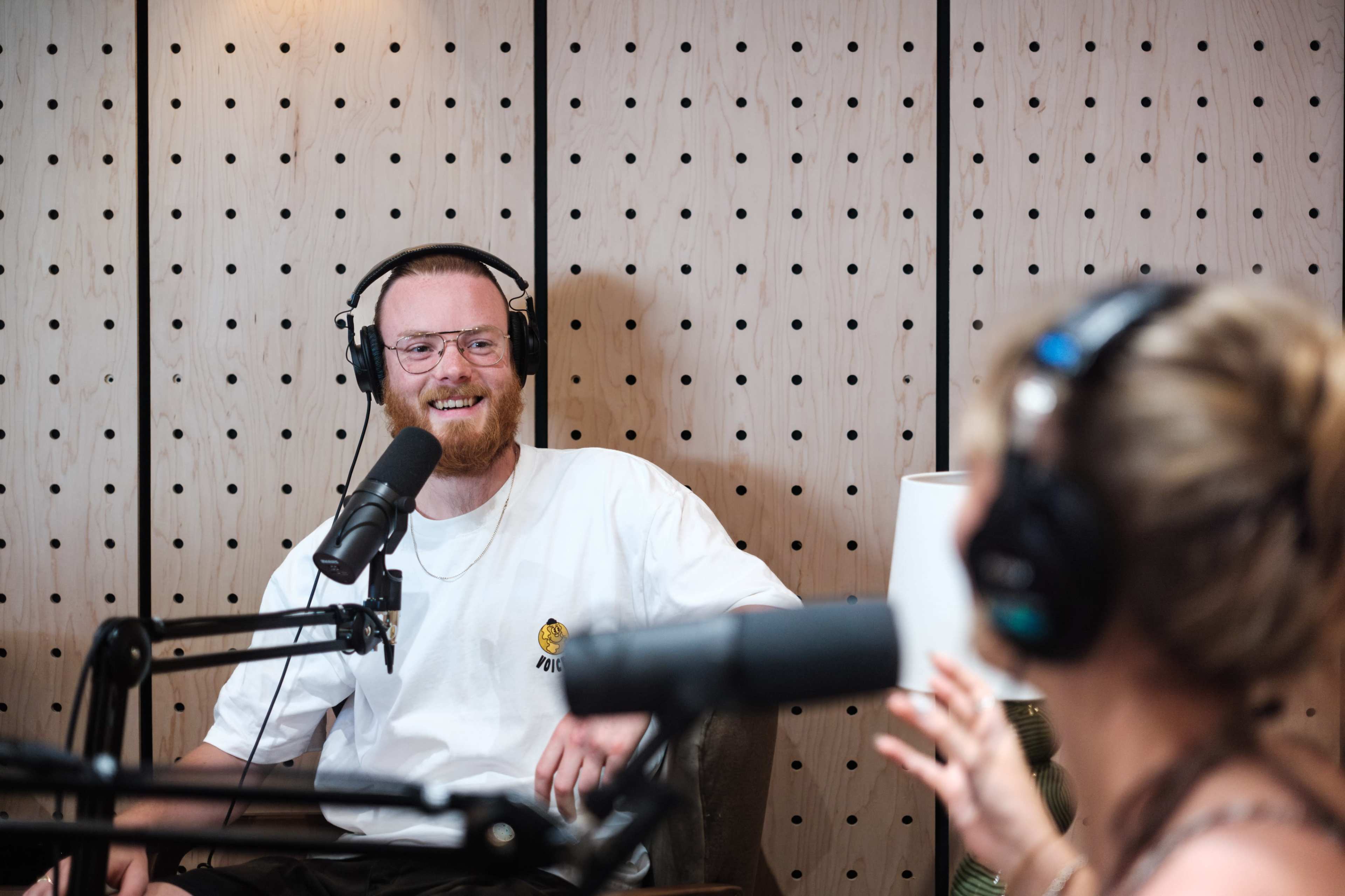 A man wearing headphones sits at a microphone during a podcast recording, smiling as he converses with a woman off-camera.