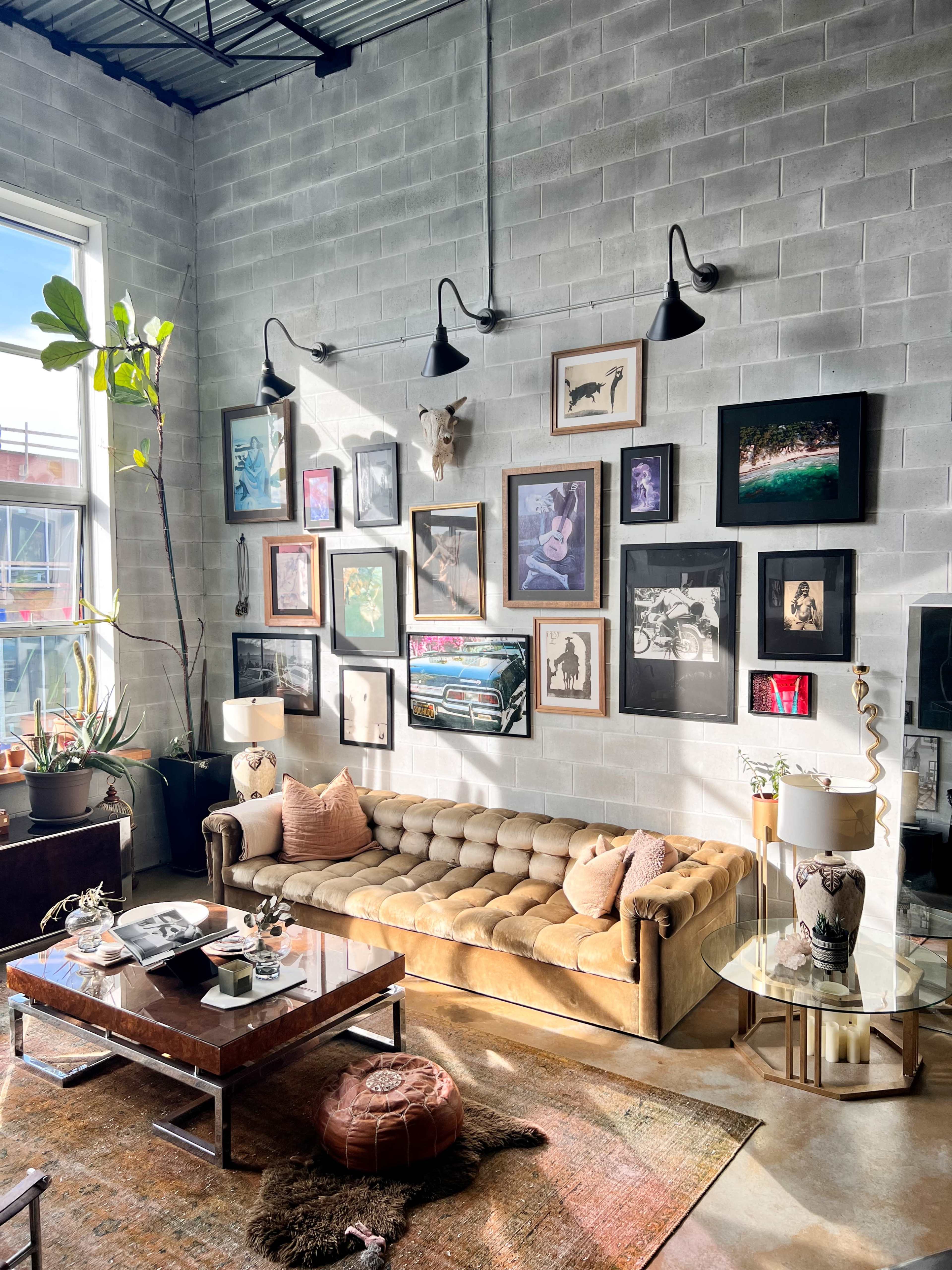 A cozy living room features a beige tufted sofa, a wall adorned with various framed artworks, and a coffee table surrounded by plants and decorative elements.